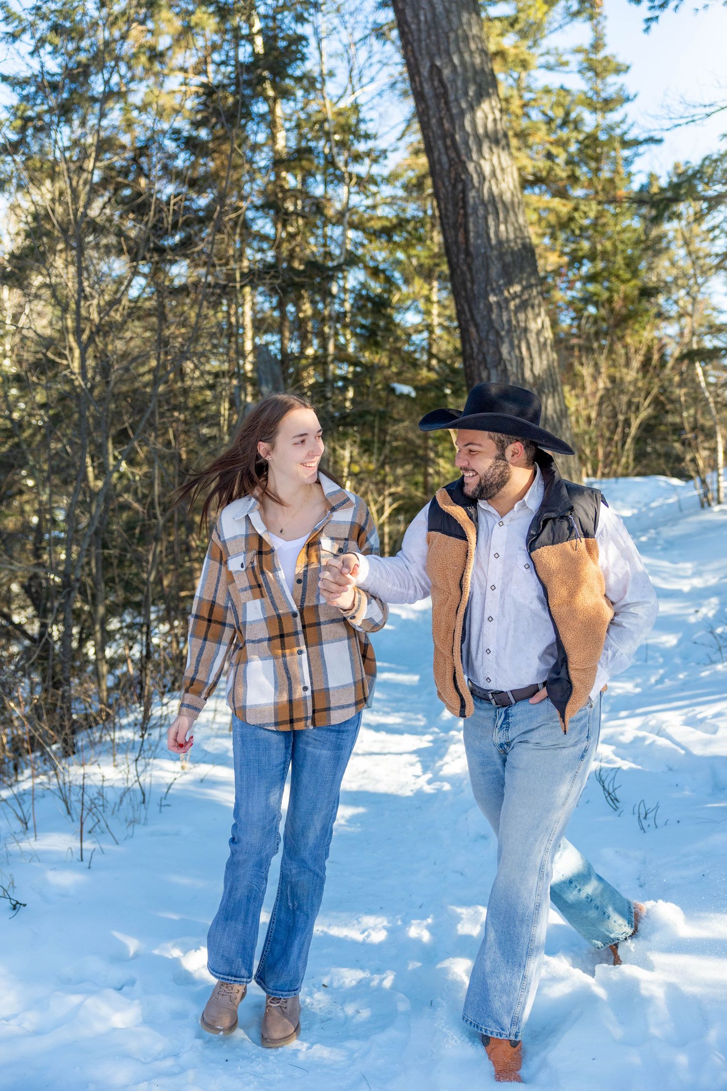 A couple playfully poses together in a snowy winter forest setting with pine trees in the background.