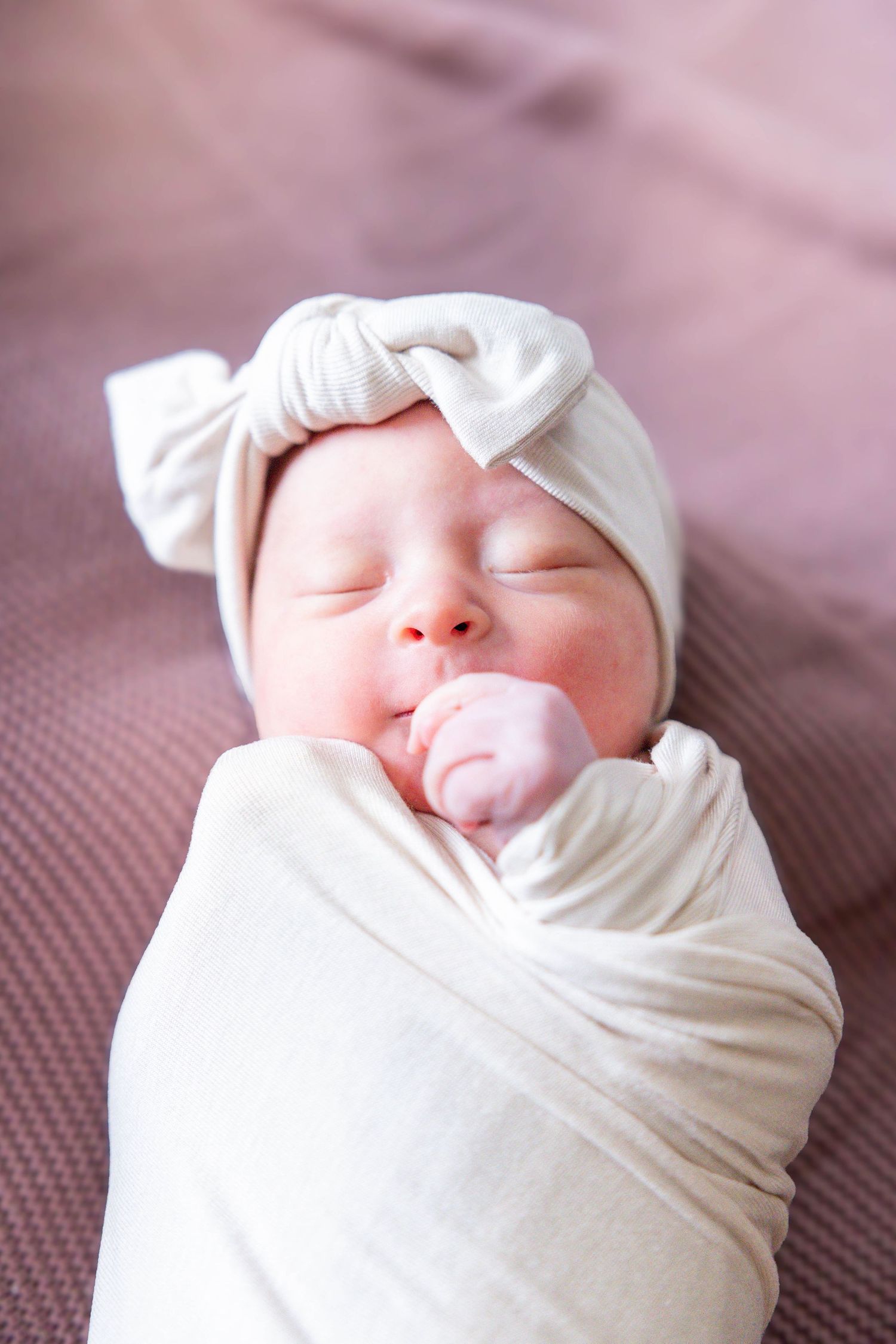 A sleeping newborn baby swaddled in soft white fabric rests peacefully against a pink textured background.