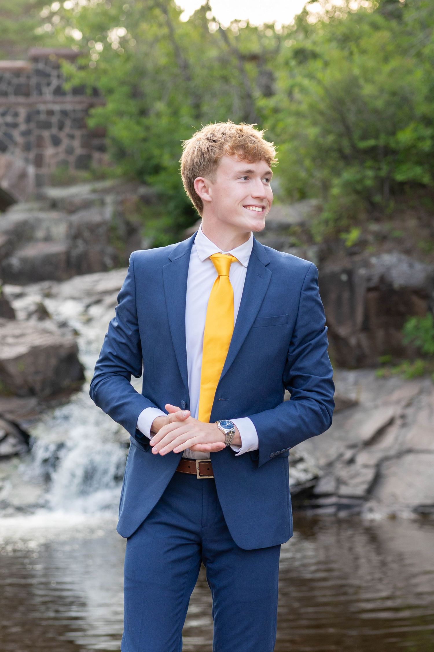 Young man in blue suit and yellow tie smiling outdoors near waterfall.