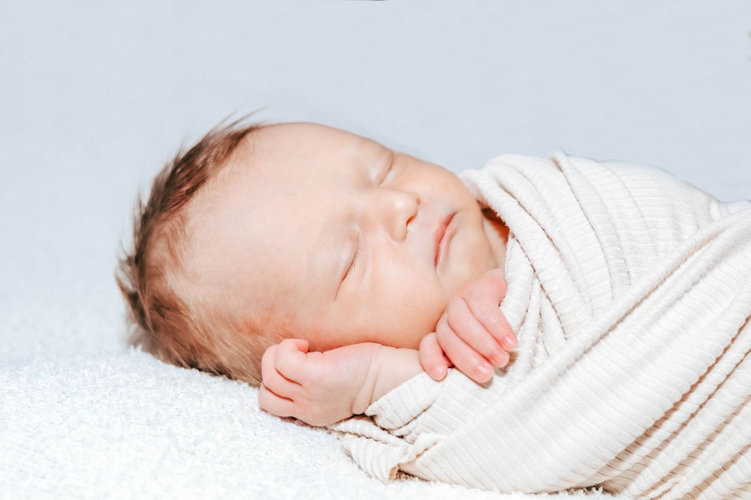 Sleeping newborn wrapped in soft white blanket lying on white bedding.
