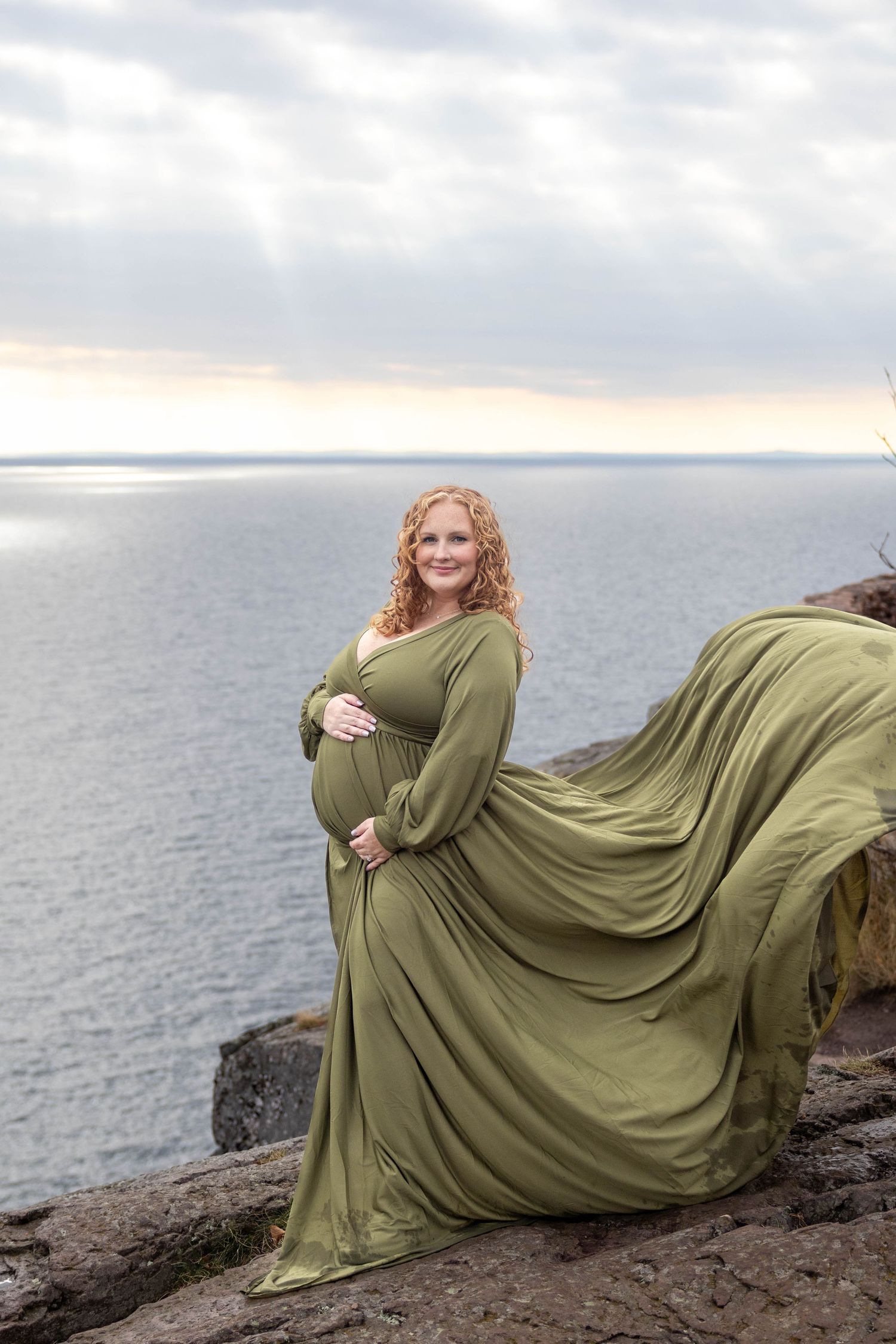 A flowing olive green dress billows dramatically against a cloudy ocean backdrop on rocky cliffs. Palisade Silver bay MN