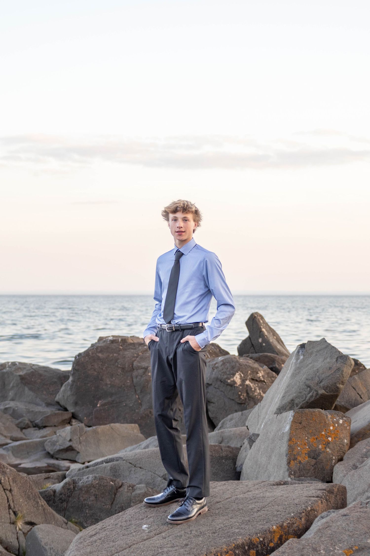 A young person in business attire stands on coastal rocks near the ocean at sunset.
