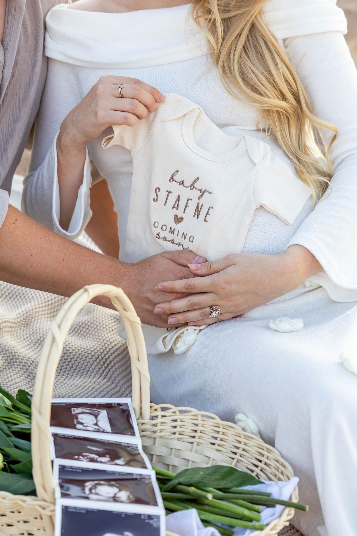 Pregnancy announcement featuring white baby onesie with text and ultrasound photos in wicker basket with greenery.