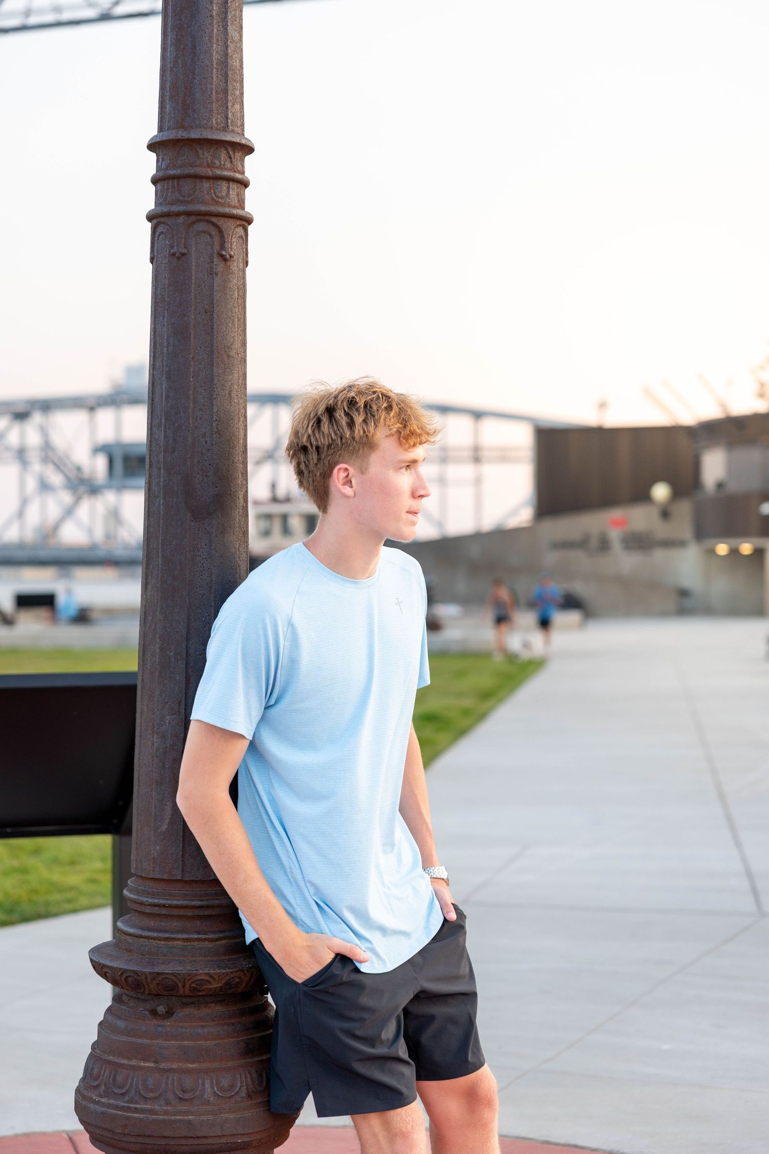 Teen boy in light blue t-shirt leans against decorative lamp post outdoors near bridge at sunset.