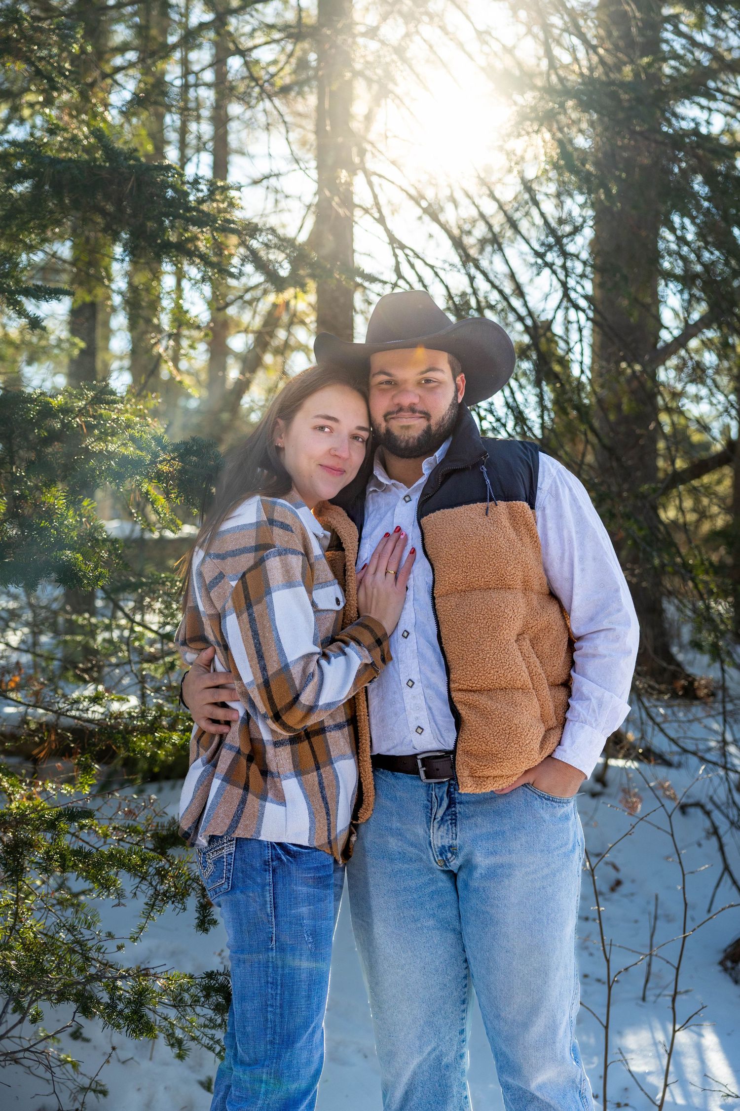 Couple in western attire and denim posing together against snowy forest backdrop during golden hour.
