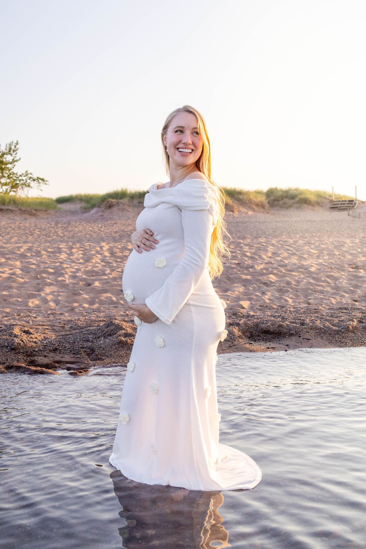 A pregnant person in a flowing white dress poses at sunset on a beach, standing in shallow water.