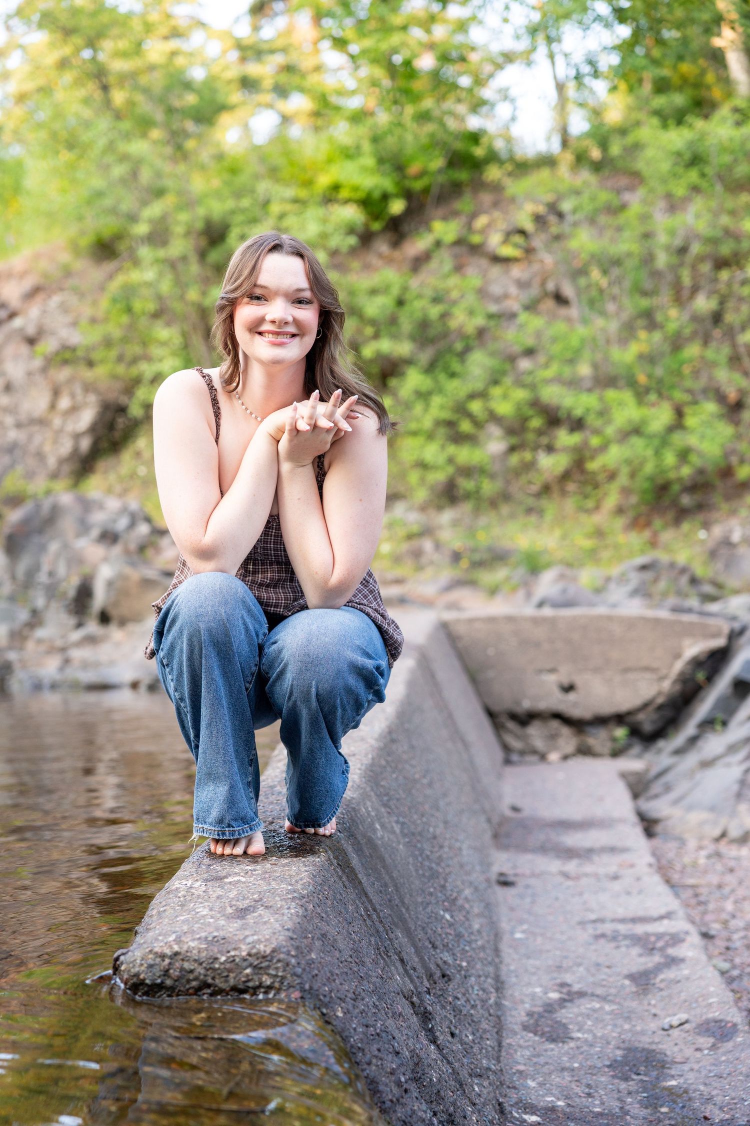 Young woman smiling, sitting on stone ledge near water, wearing jeans and tank top, surrounded by lush green trees.
