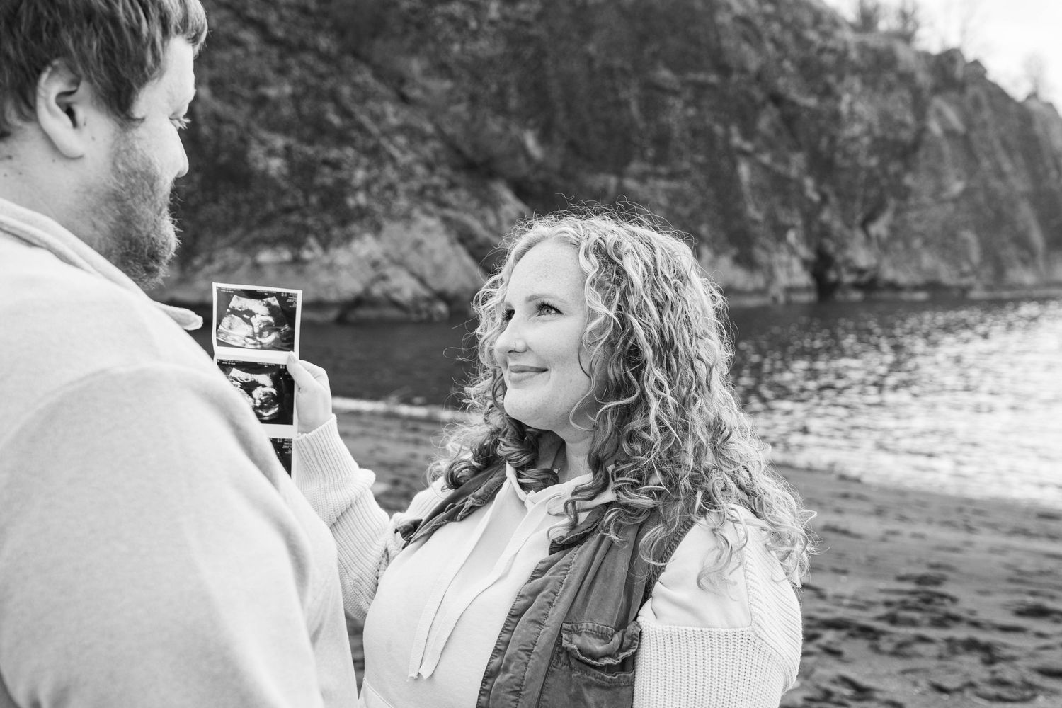 A couple shares an intimate black and white moment while looking at an ultrasound photo by the water.