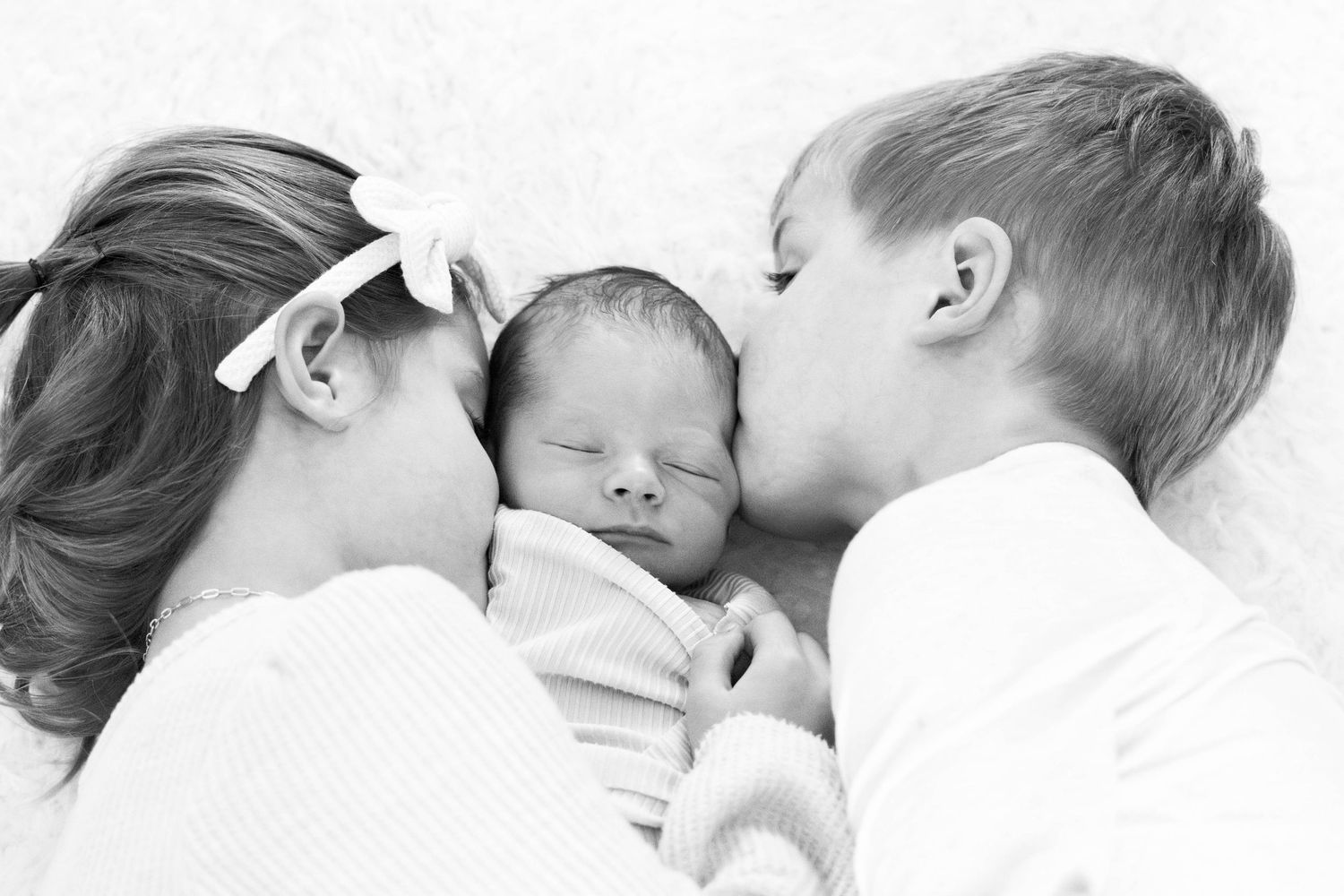 Sweet black and white portrait of siblings kissing newborn baby.