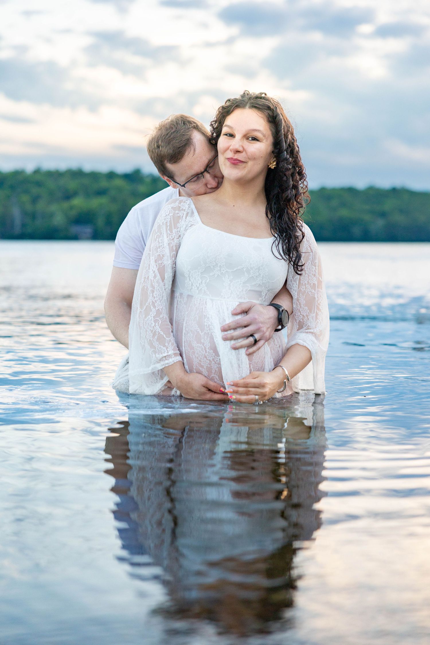 Pregnant couple standing in a lake, man kissing woman's cheek while holding her baby bump, wearing white outfits.