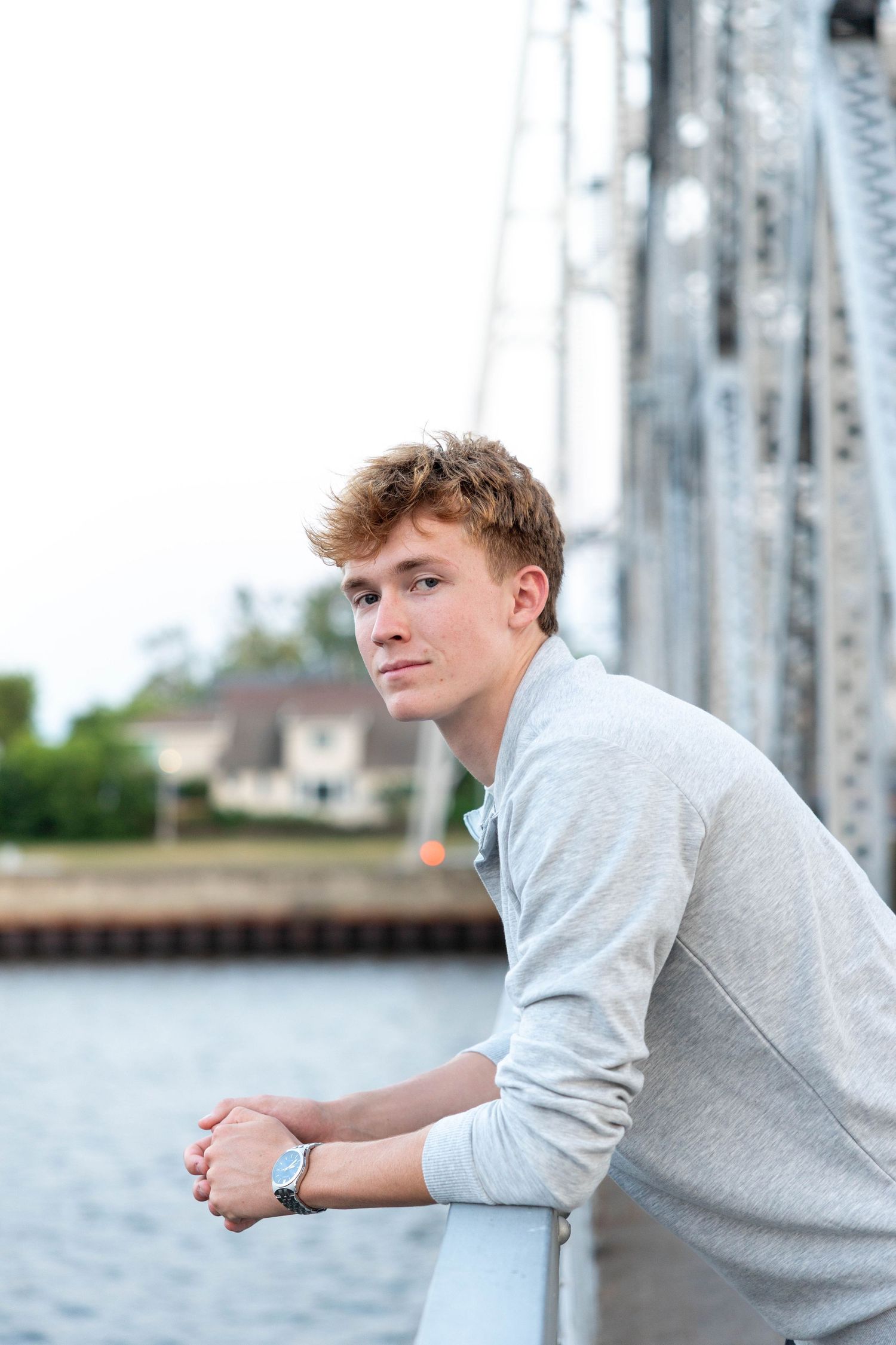 Young man in gray sweatshirt leaning on bridge railing near waterfront, wearing a watch, looking thoughtfully to the side.
