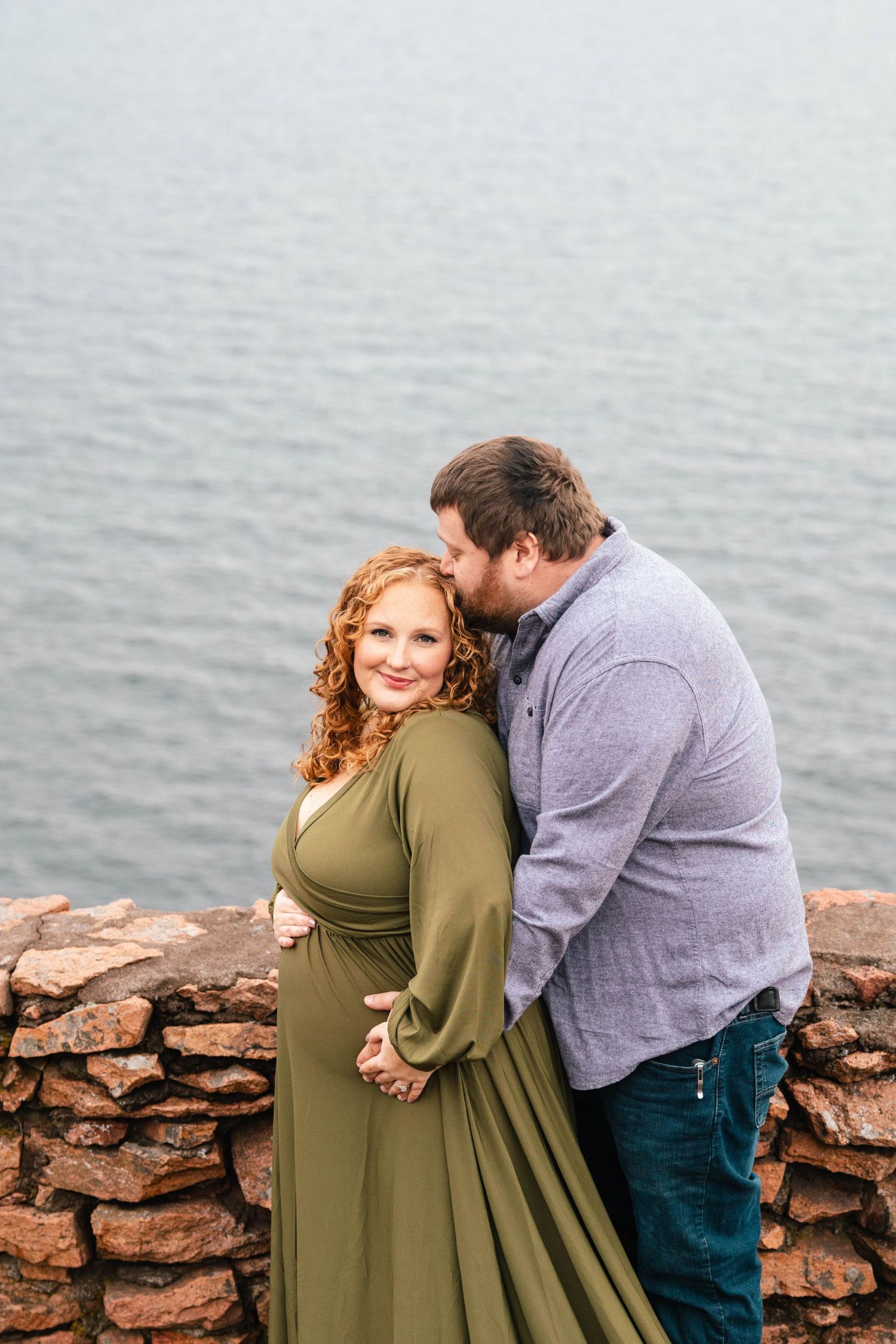 Romantic maternity photo of couple embracing by stone wall overlooking ocean.