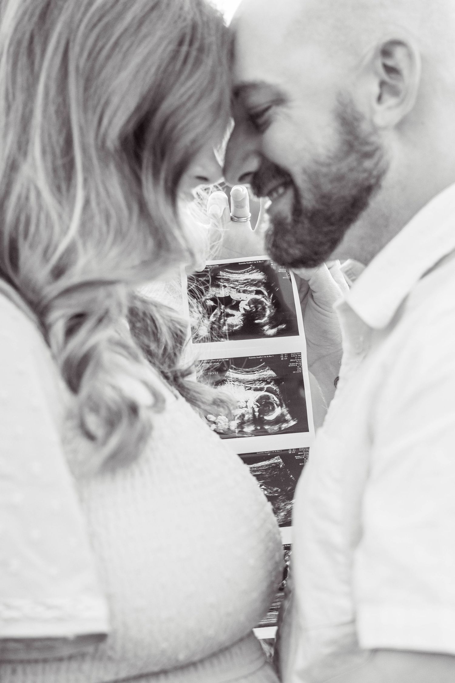 A couple shares an intimate moment while holding ultrasound photos of their expected baby in a black and white portrait.