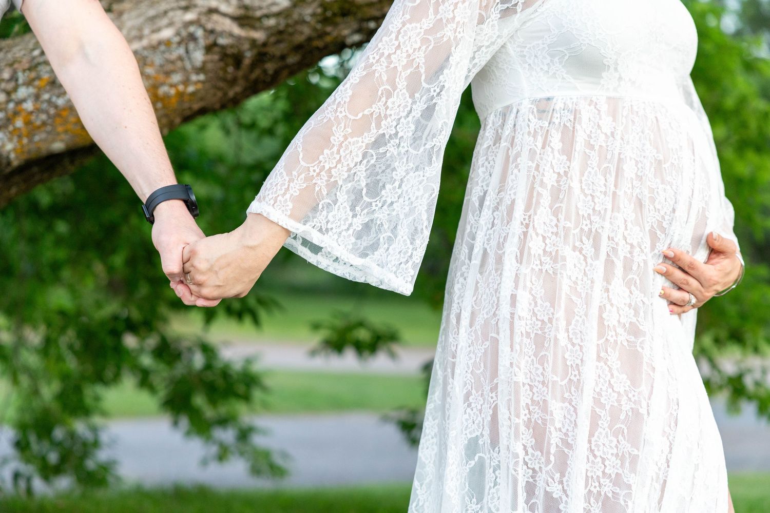 Pregnant woman in white lace dress holds partner's hand outdoors, maternity photo shoot in green park setting.