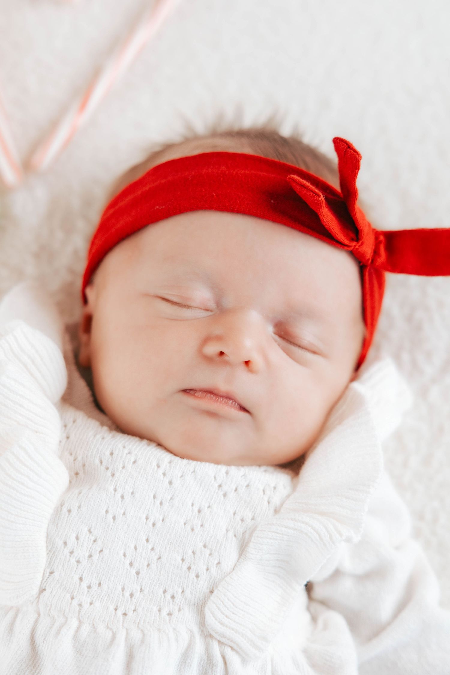 Sweet sleeping baby wearing white outfit and red bow headband on white blanket.