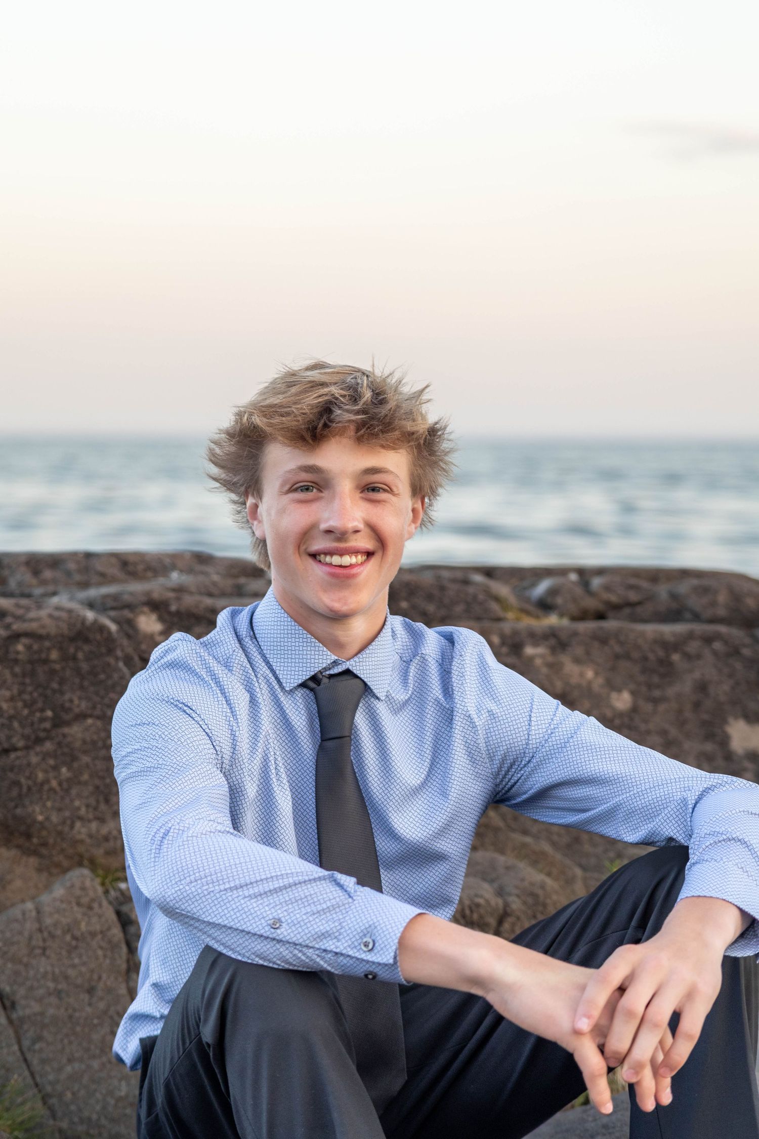 Professional portrait against rocky coastal backdrop showing person in light blue dress shirt and dark tie at sunset.