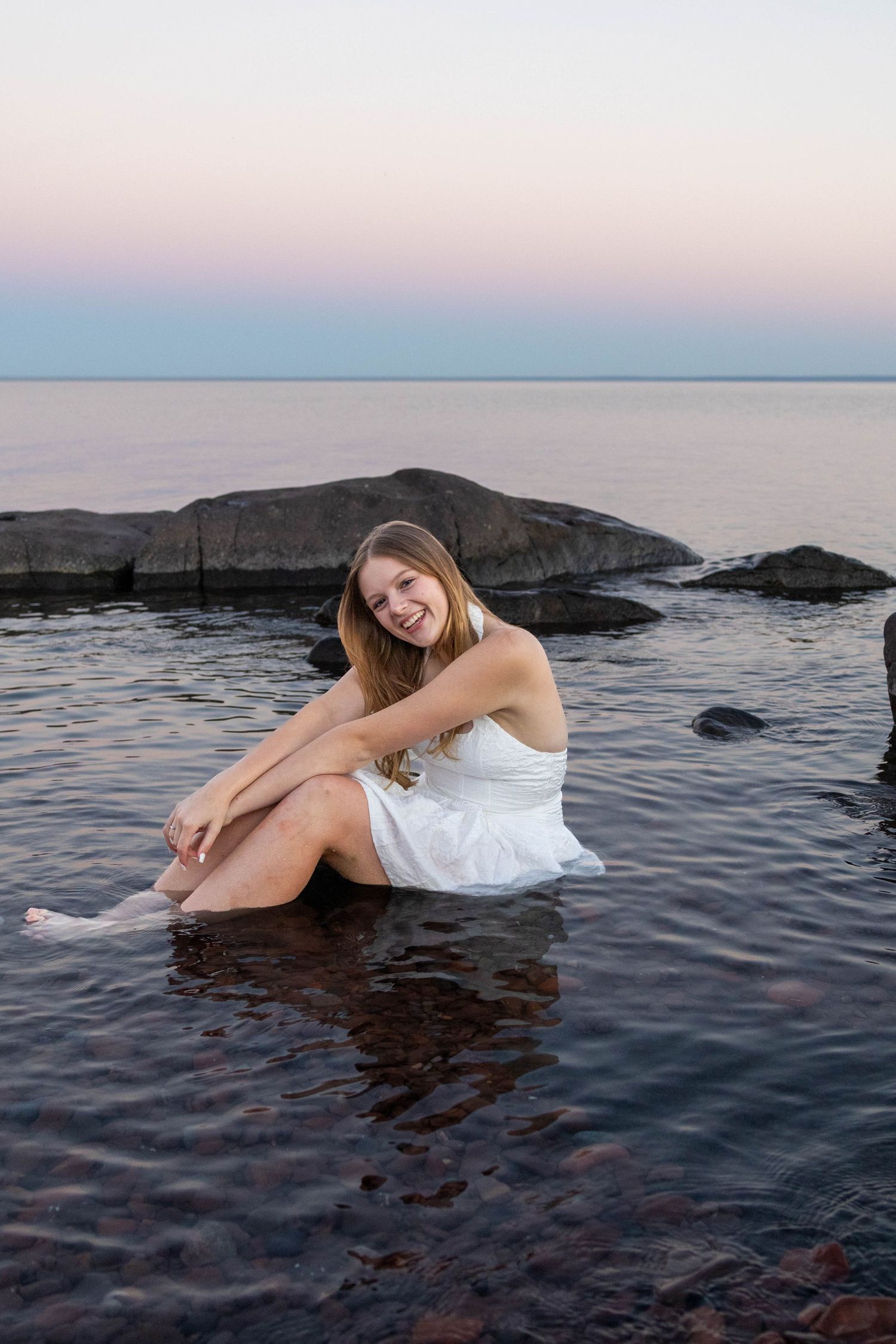 A person in a white dress poses artistically in shallow ocean water during sunset with rocky coastline in background.