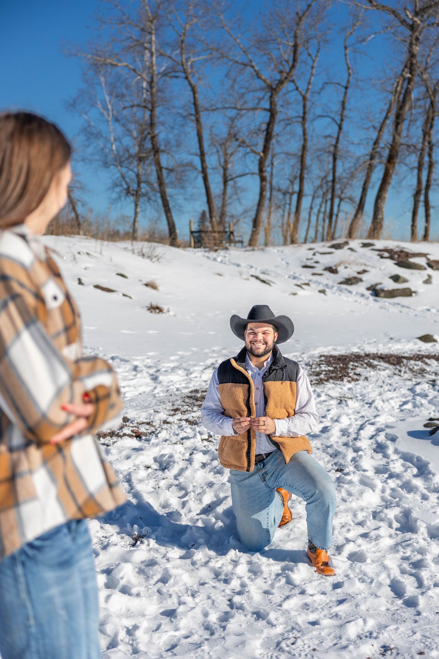 Couple taking winter engagement photos in a snowy landscape with bare trees and bright sunlight.