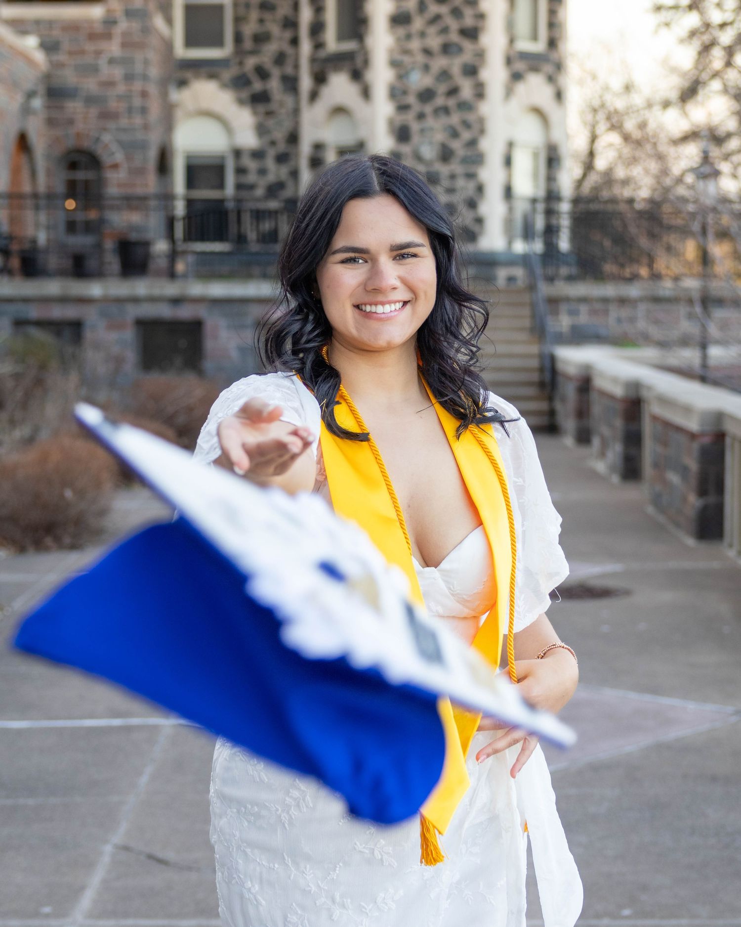 A graduate celebrates in a yellow academic stole and white dress outside a stone church building on a sunny day.