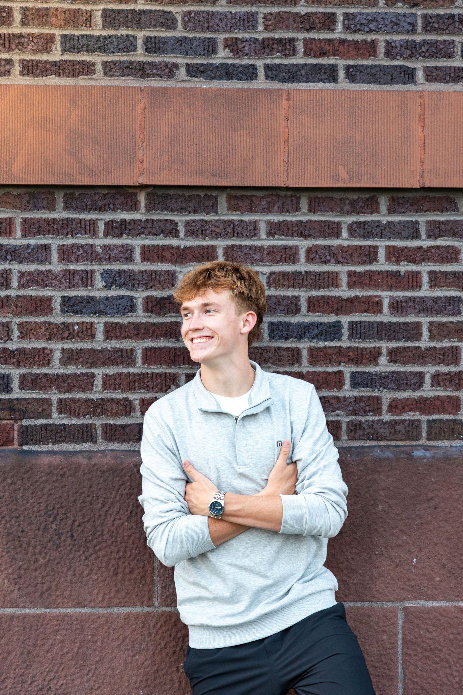 Smiling teenage boy in gray sweatshirt with arms crossed leaning against a brick wall outdoors.