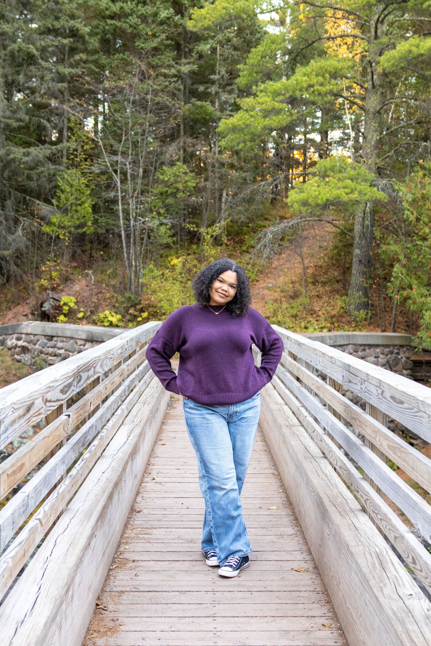 Person in purple sweater and jeans stands on wooden boardwalk surrounded by autumn forest scenery.