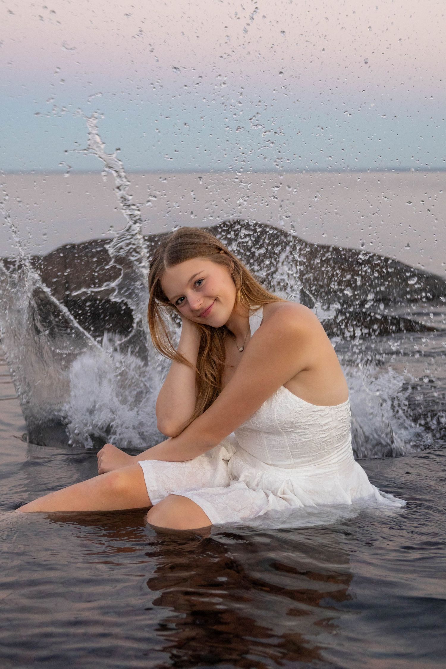 A woman in a white dress poses gracefully in ocean waves during a beach photoshoot at sunset.