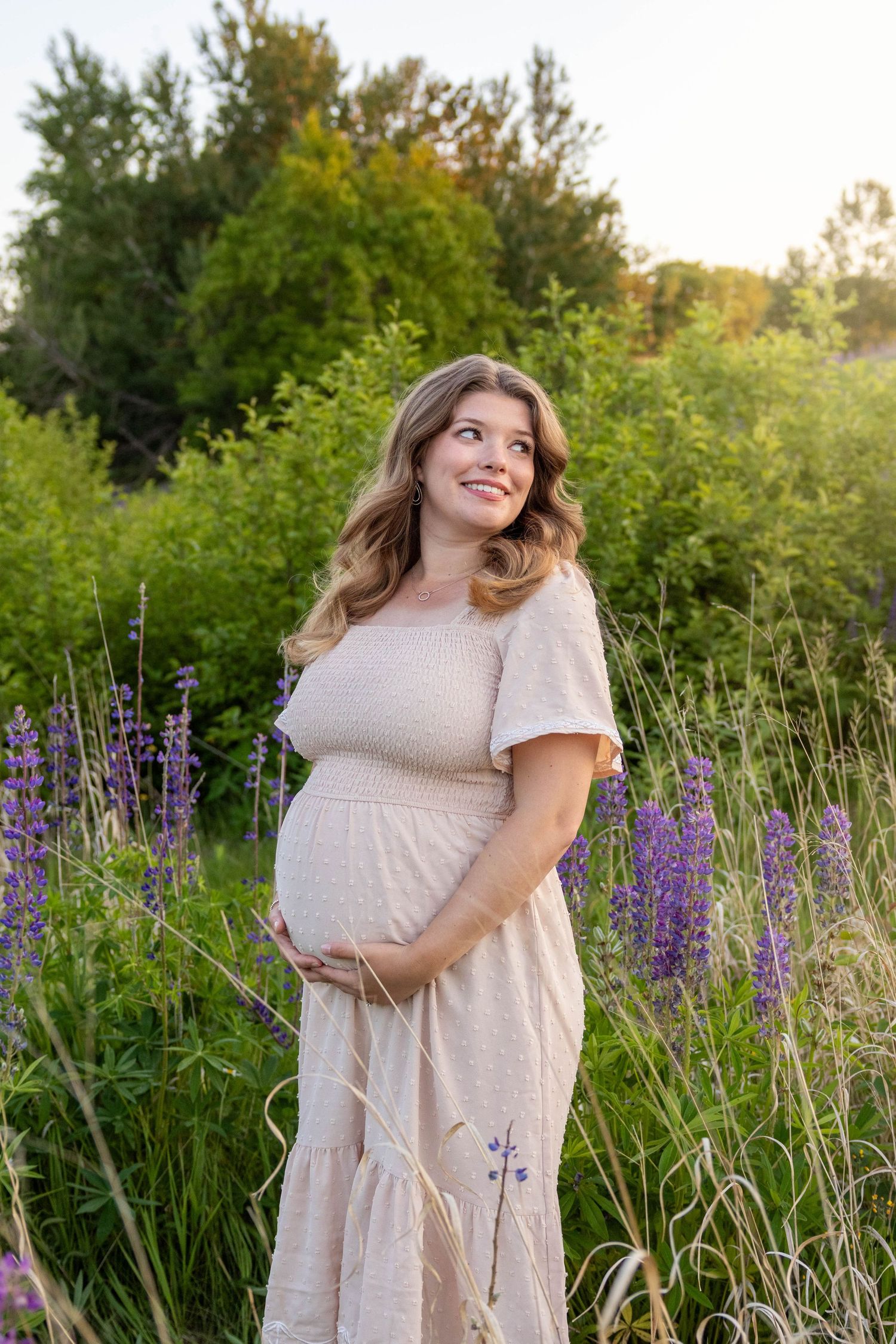 A person in a cream dress poses in a field of purple lupine flowers during golden hour.