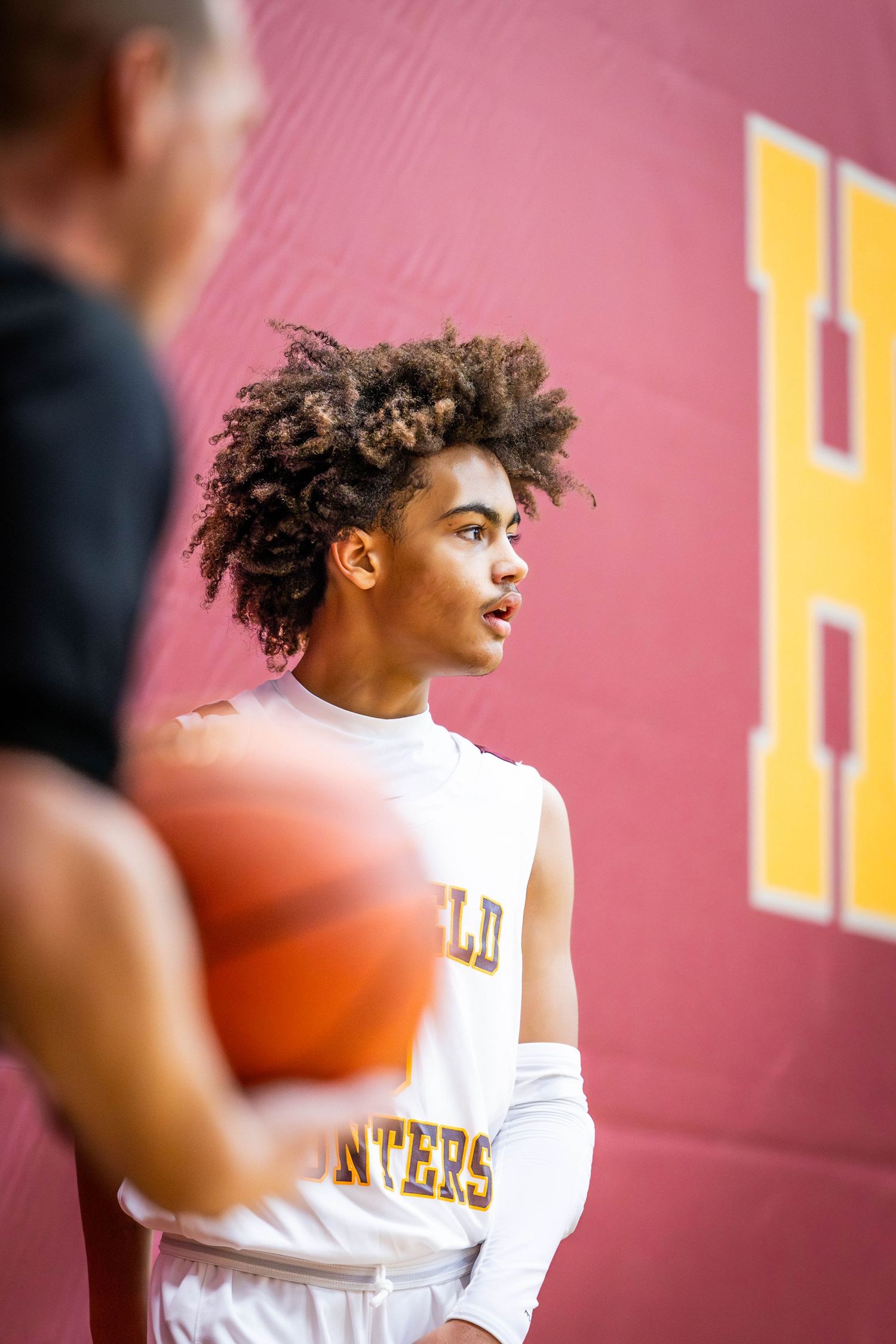 Basketball player in white jersey with curly hair shown in profile against pink background during practice.