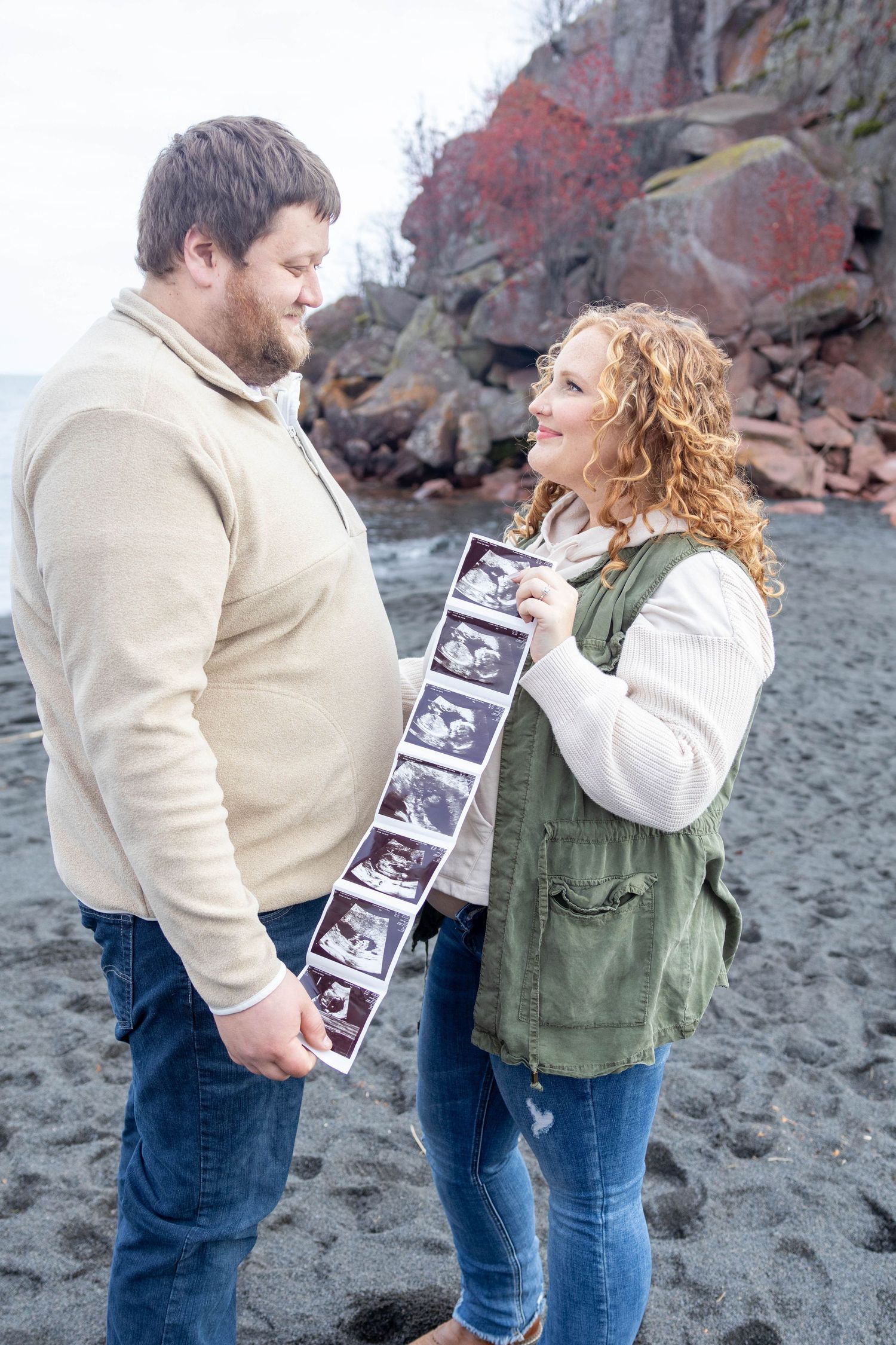 A couple shares a joyful moment looking at ultrasound photos on a rocky beach.