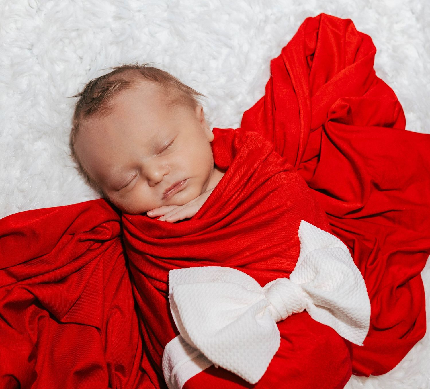 Peaceful newborn swaddled in red fabric with white bow sleeping on textured background.