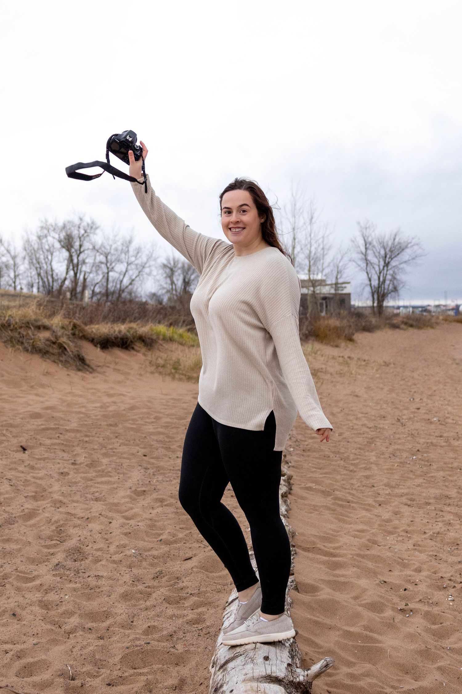 Person in cream sweater joyfully poses with camera on driftwood at beach.