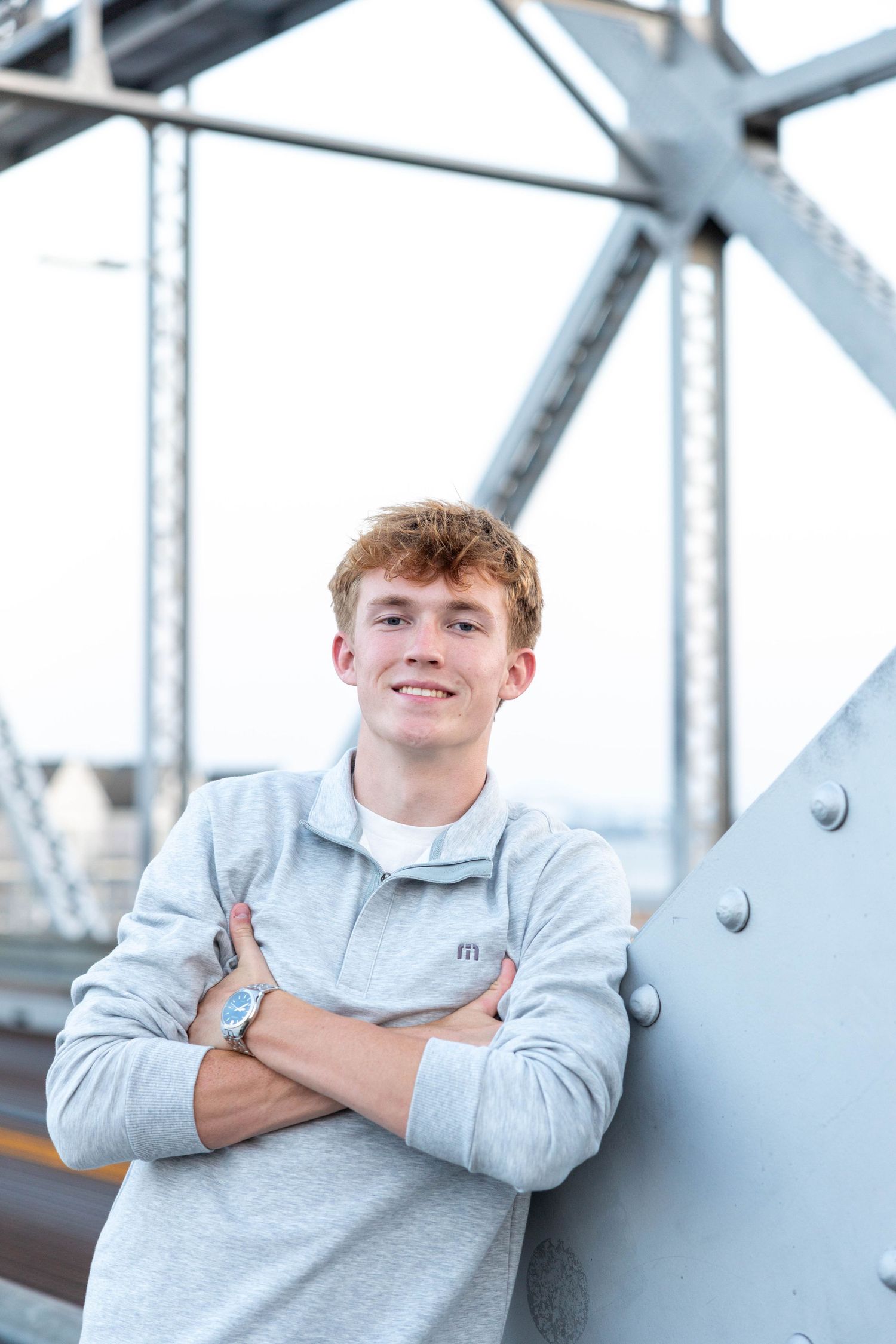Young man with red hair in gray sweatshirt smiles with arms crossed, leaning against a steel bridge structure.