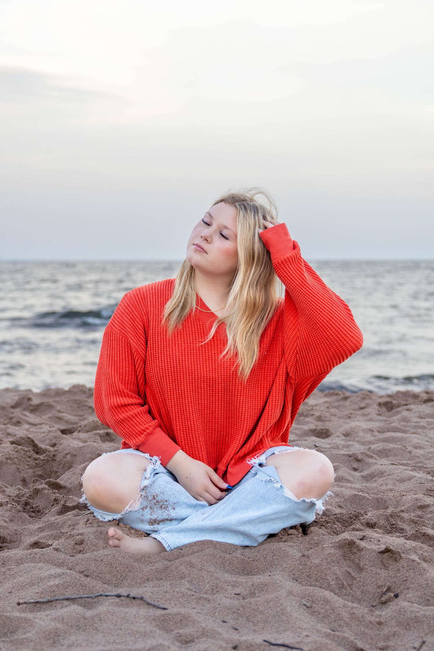 A person in a bright red sweater and light jeans sits on a sandy beach by the ocean at sunset.