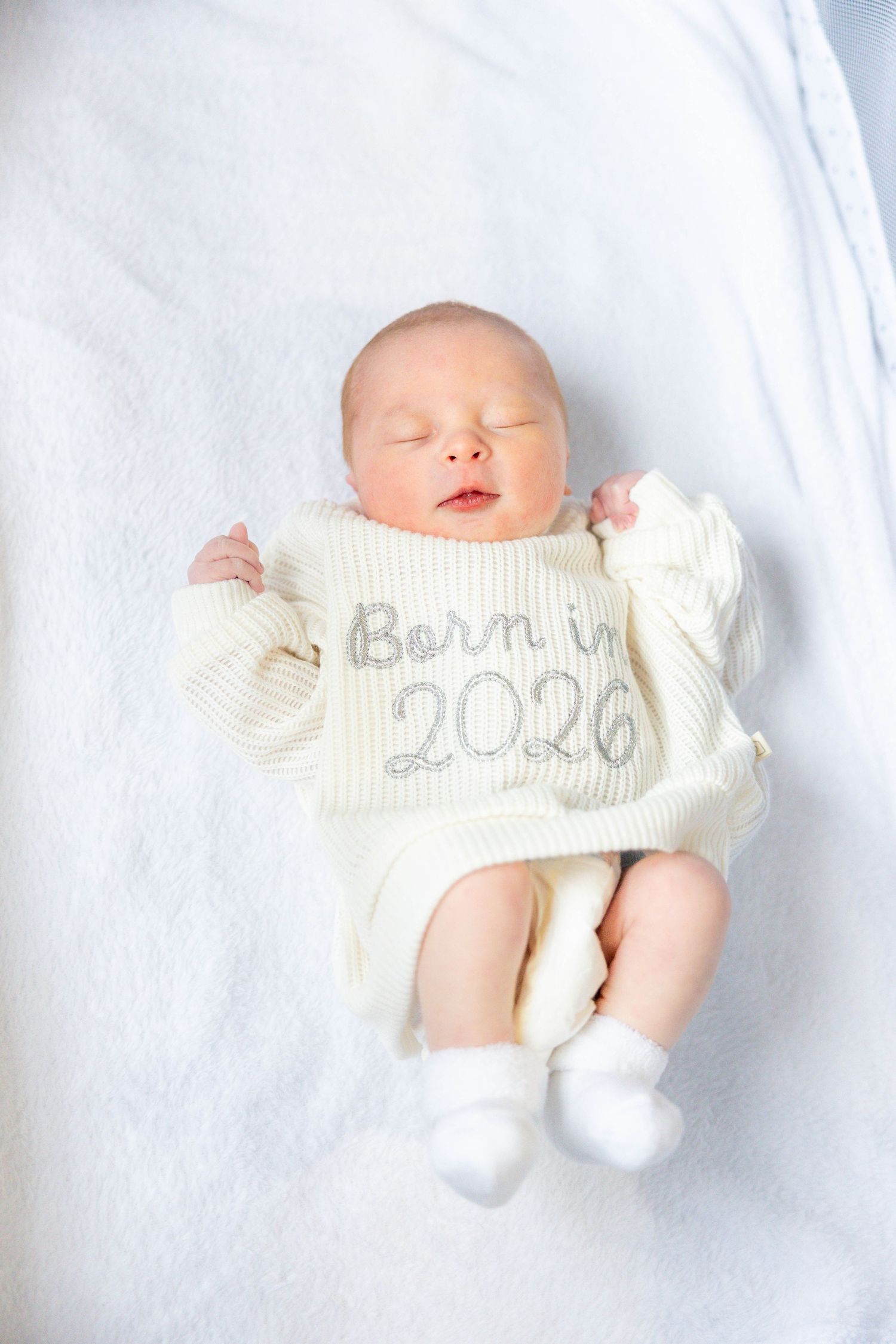 A sleeping newborn baby wearing white socks lies on white bedding with 2020 written on their clothing.