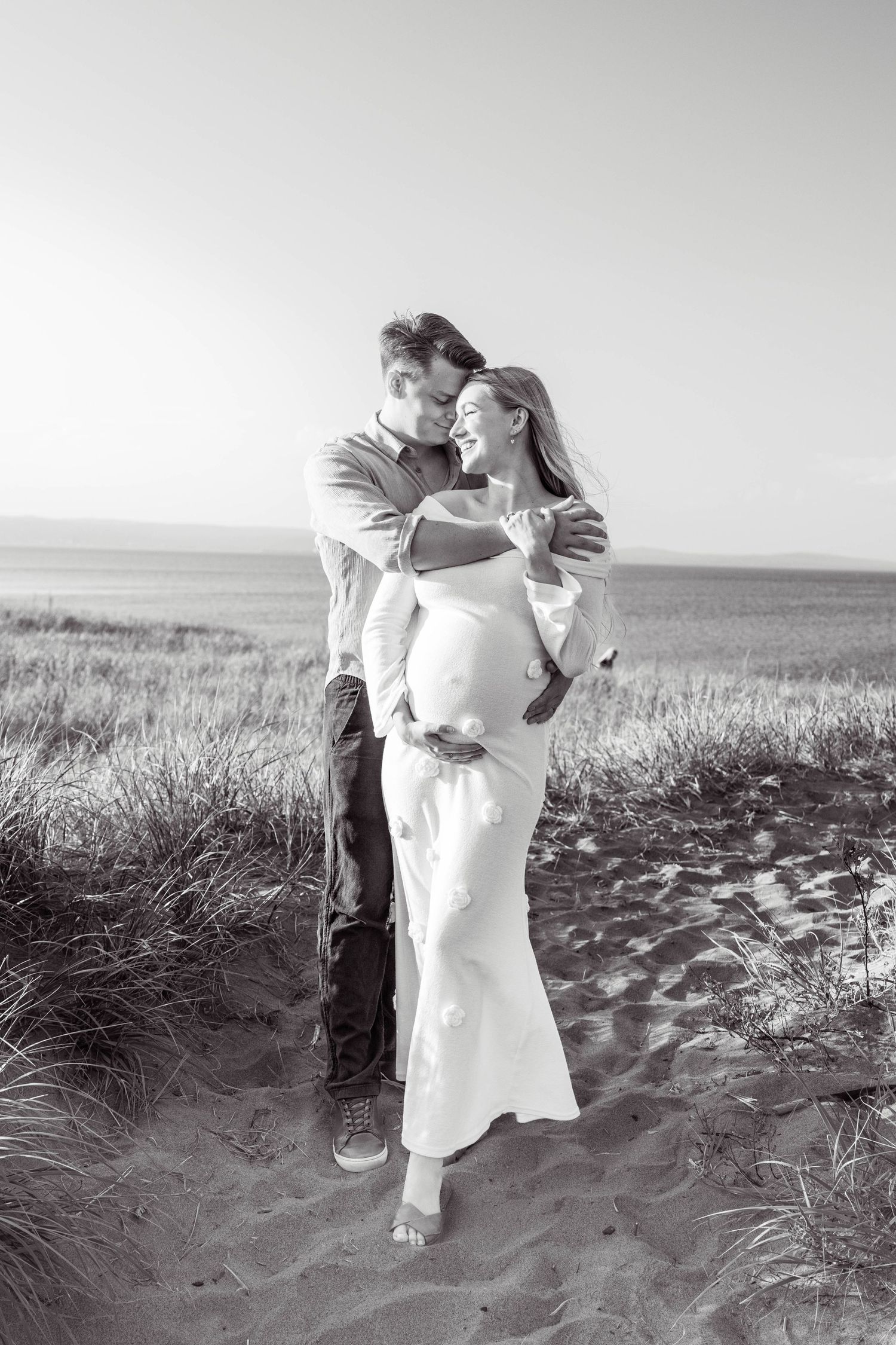 A romantic black and white sequence shows a moment of joy between two people embracing on a beach by tall grass.
