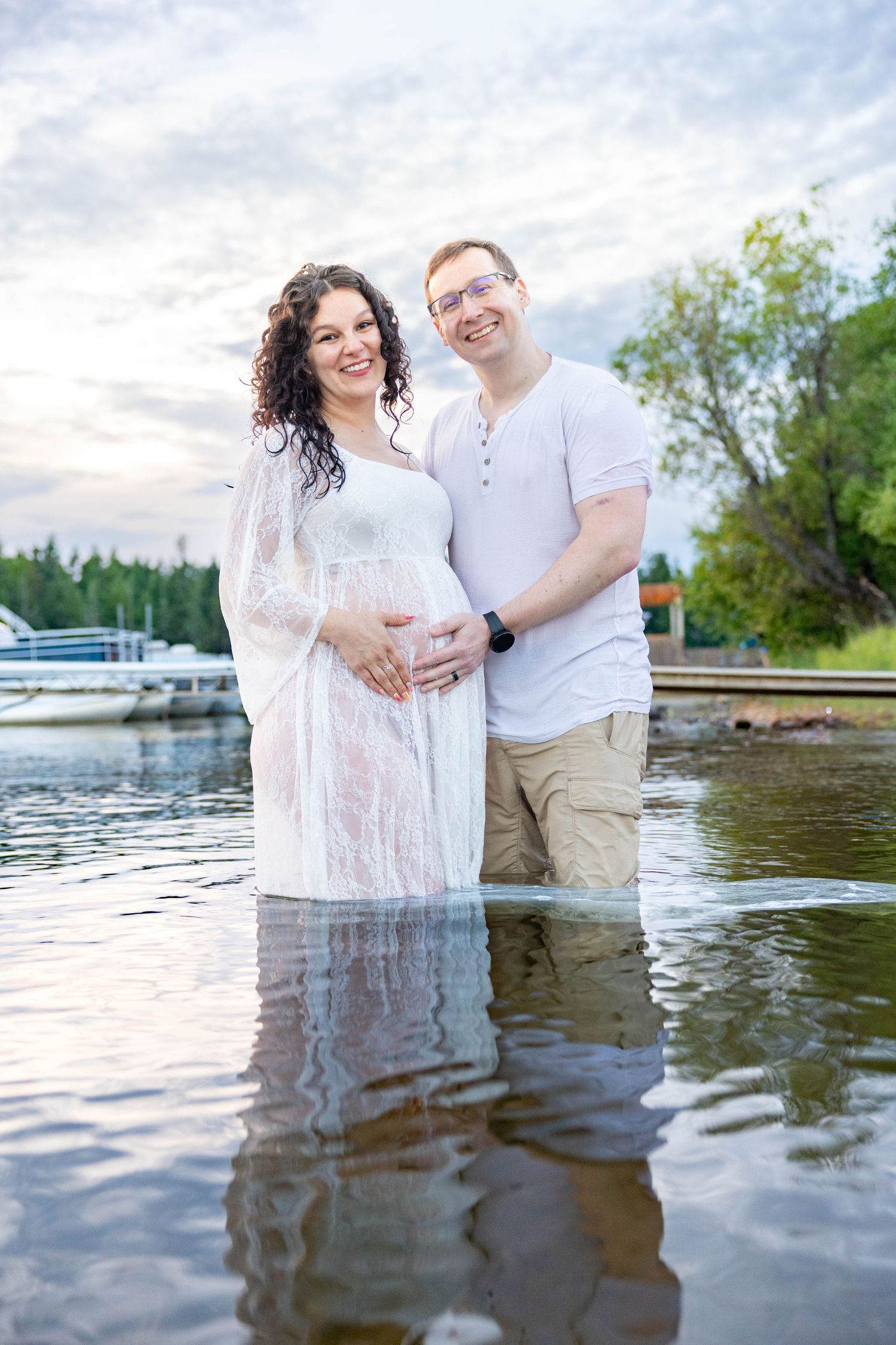Smiling pregnant couple standing in shallow water, woman in white dress with hands on baby bump, man in white shirt and khaki shorts.