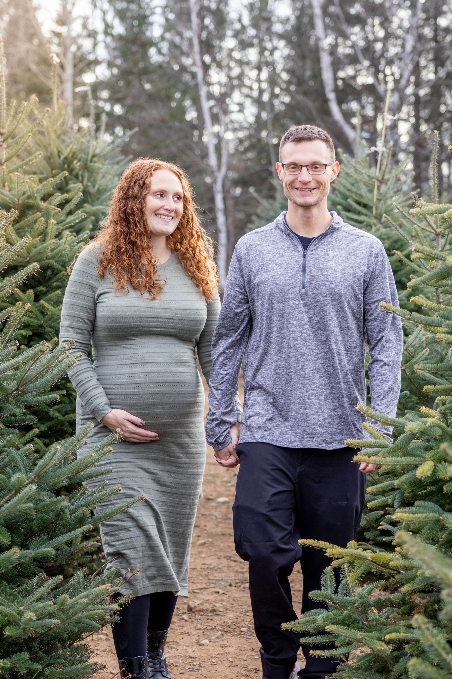 Expecting couple poses together at a Christmas tree farm wearing coordinating gray outfits.