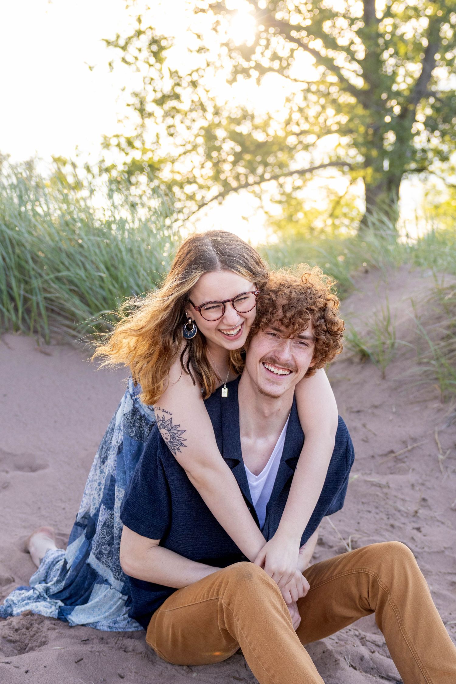 A happy couple sits together on sandy beach dunes during golden hour with sunlit trees in background.