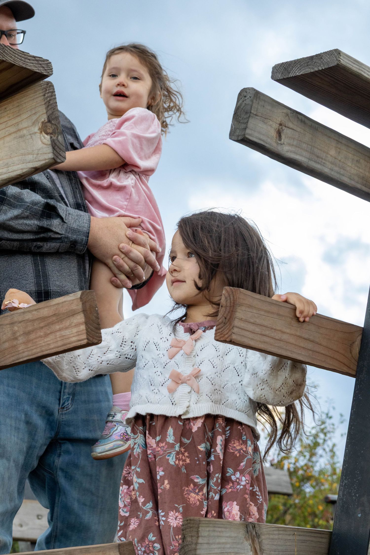 A series of outdoor photos showing a child in a pink top and floral skirt being lifted up near wooden beams.