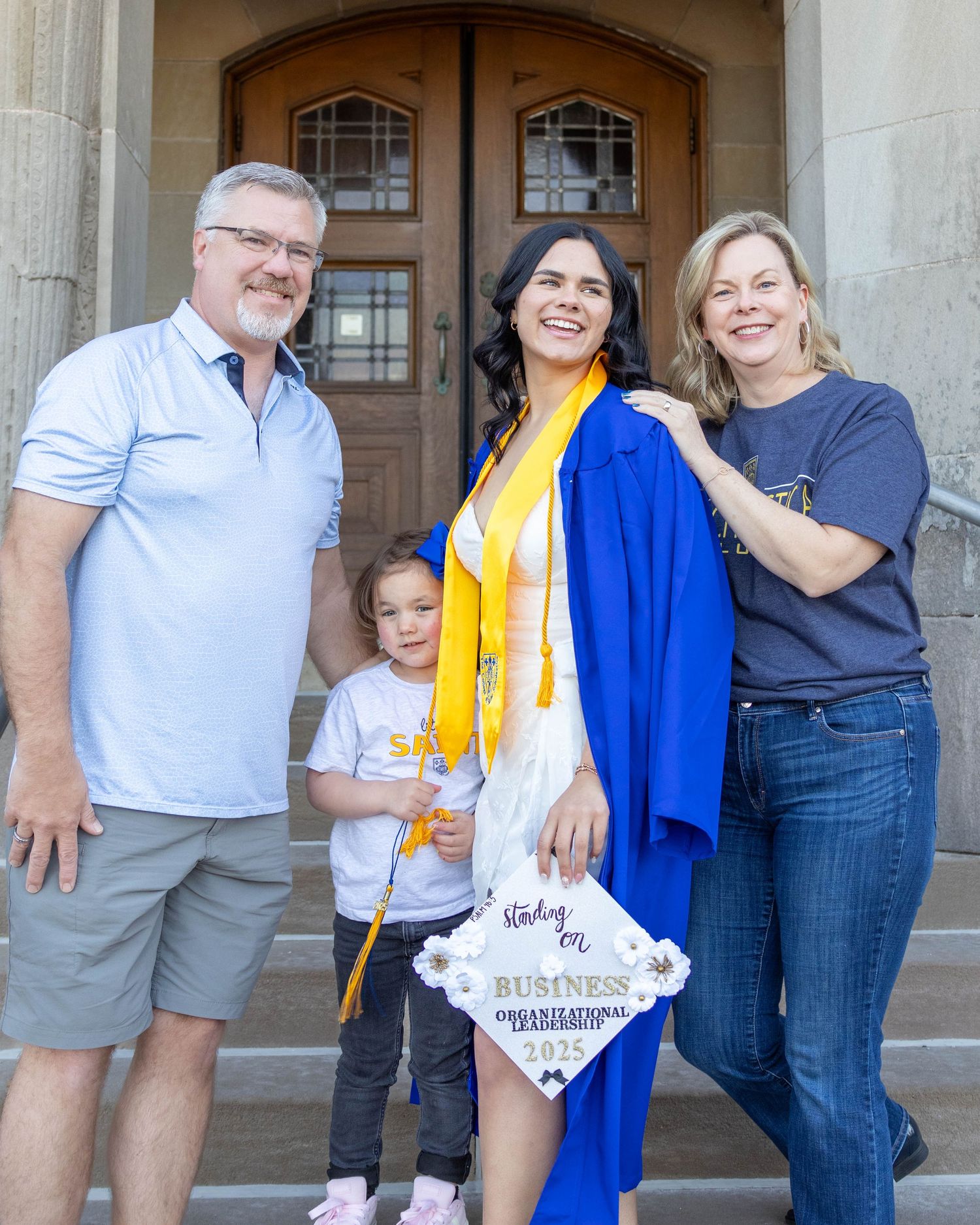 Graduate in blue and gold regalia celebrates with family on stone steps of academic building.