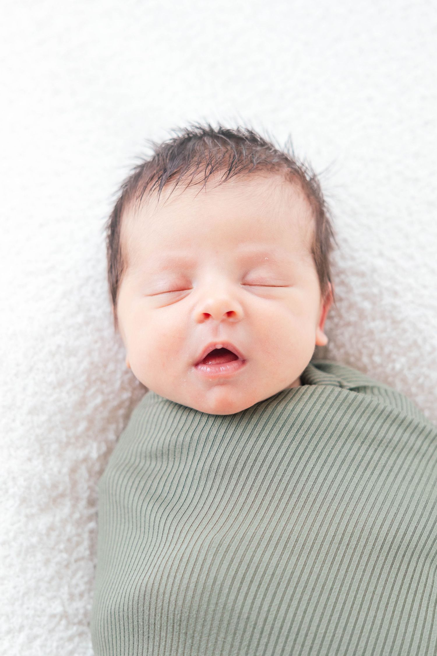 A peaceful sleeping newborn baby wrapped in a sage green ribbed swaddle against white background.