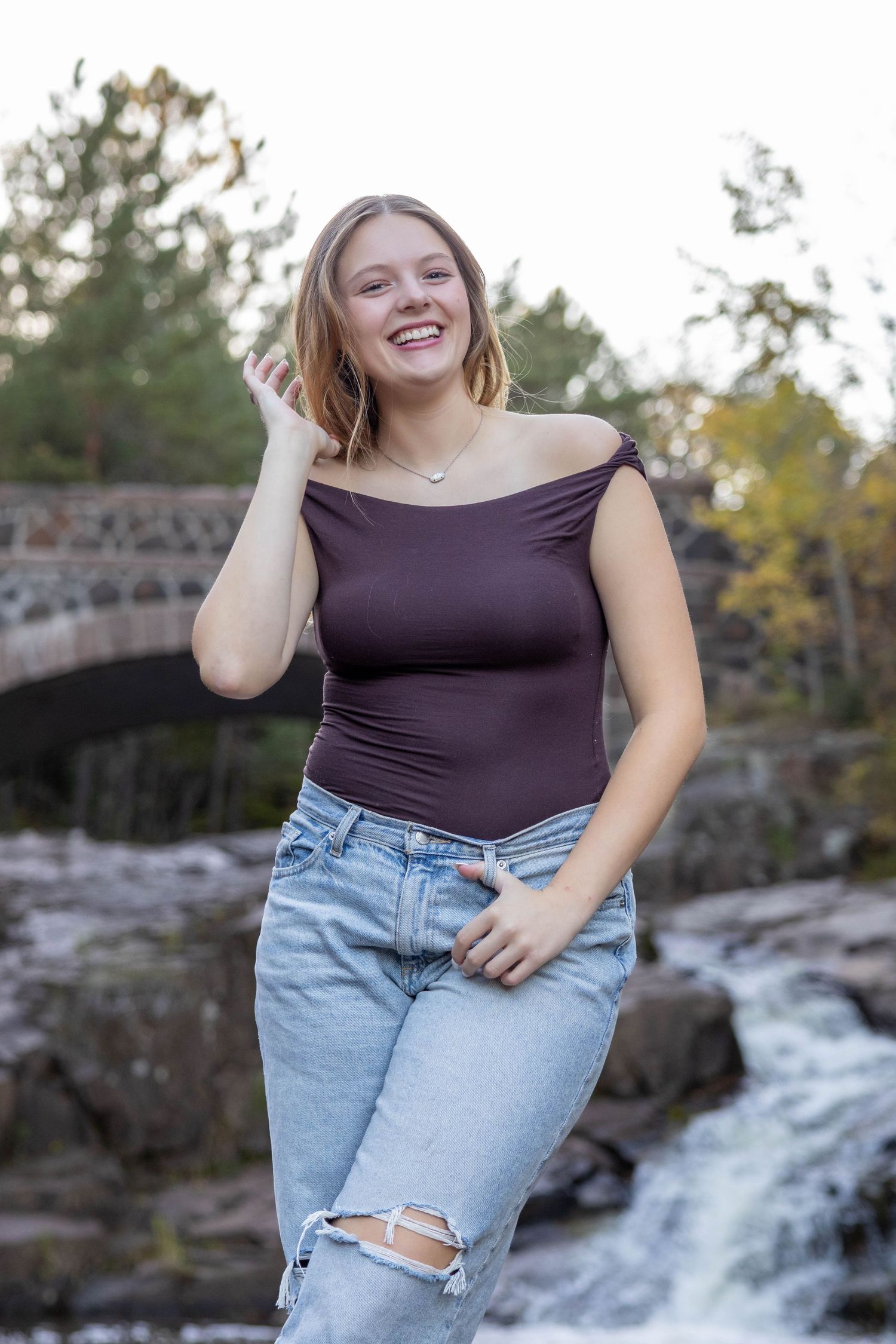 A person in a dark top and ripped jeans poses cheerfully by a scenic waterfall and stone bridge on a sunny day.