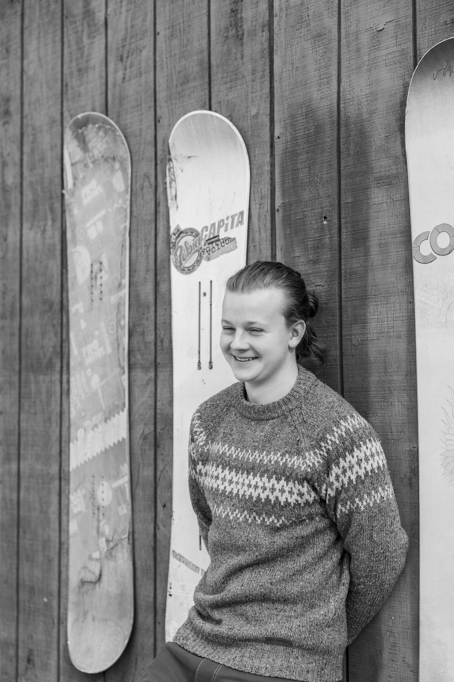 Black and white photo of a smiling young person in a Nordic sweater leaning against wooden wall with Capita snowboards.