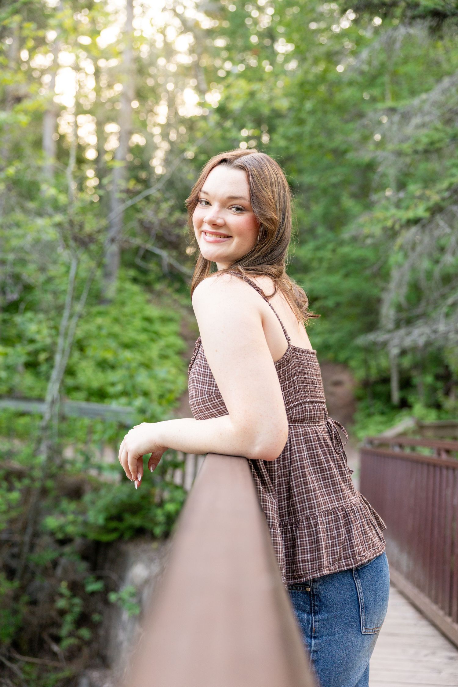Smiling young woman in plaid tank top leans on bridge railing surrounded by lush green forest trees.