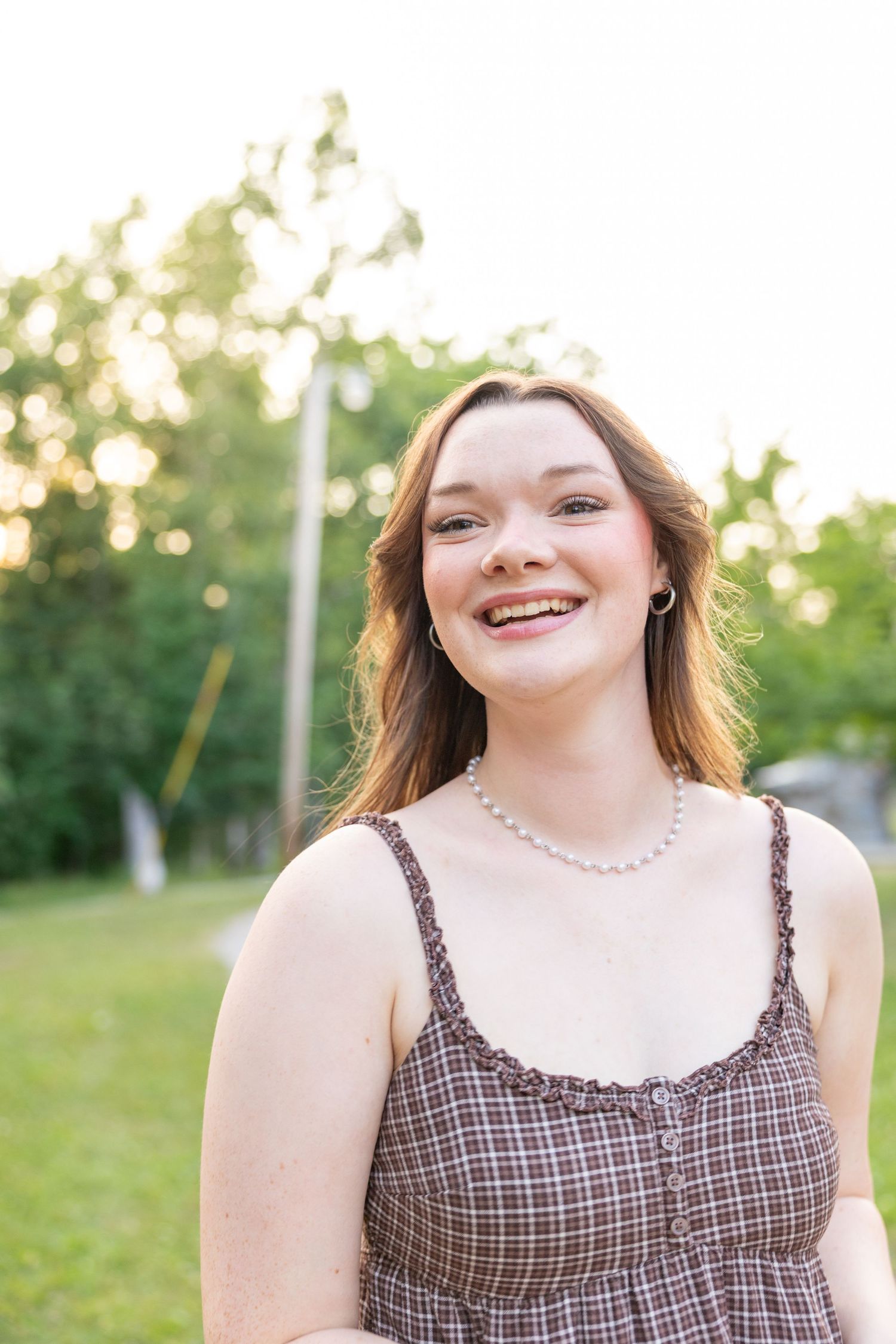 Smiling young woman in plaid sundress and pearl necklace outdoors with green trees in golden hour light.