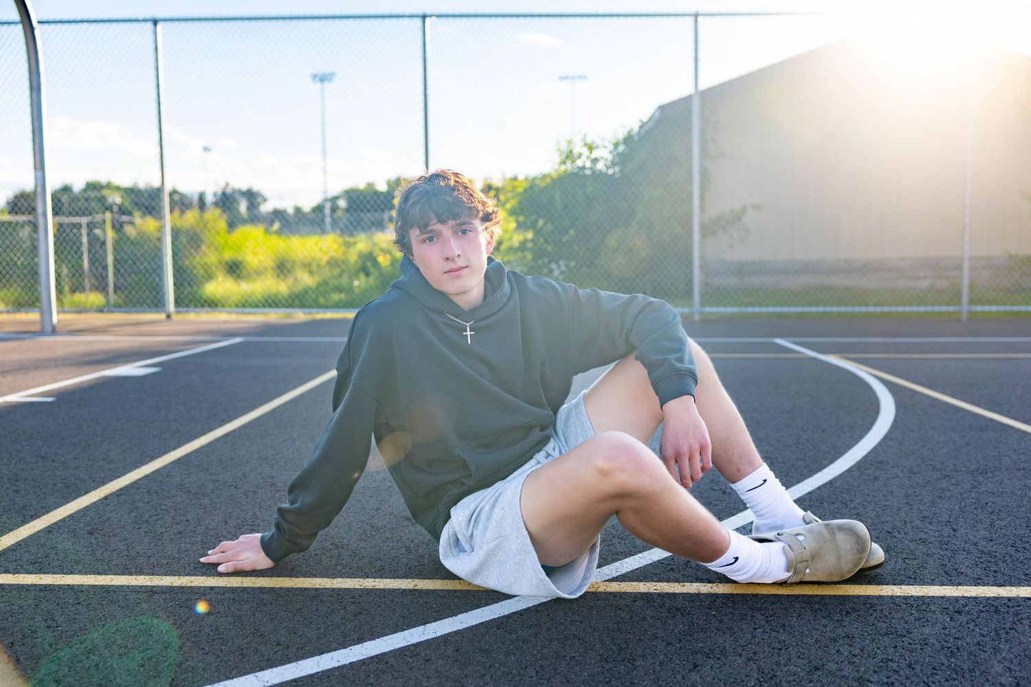 Teen boy in gray hoodie and white shorts sits on a running track outdoors in golden hour sunlight.