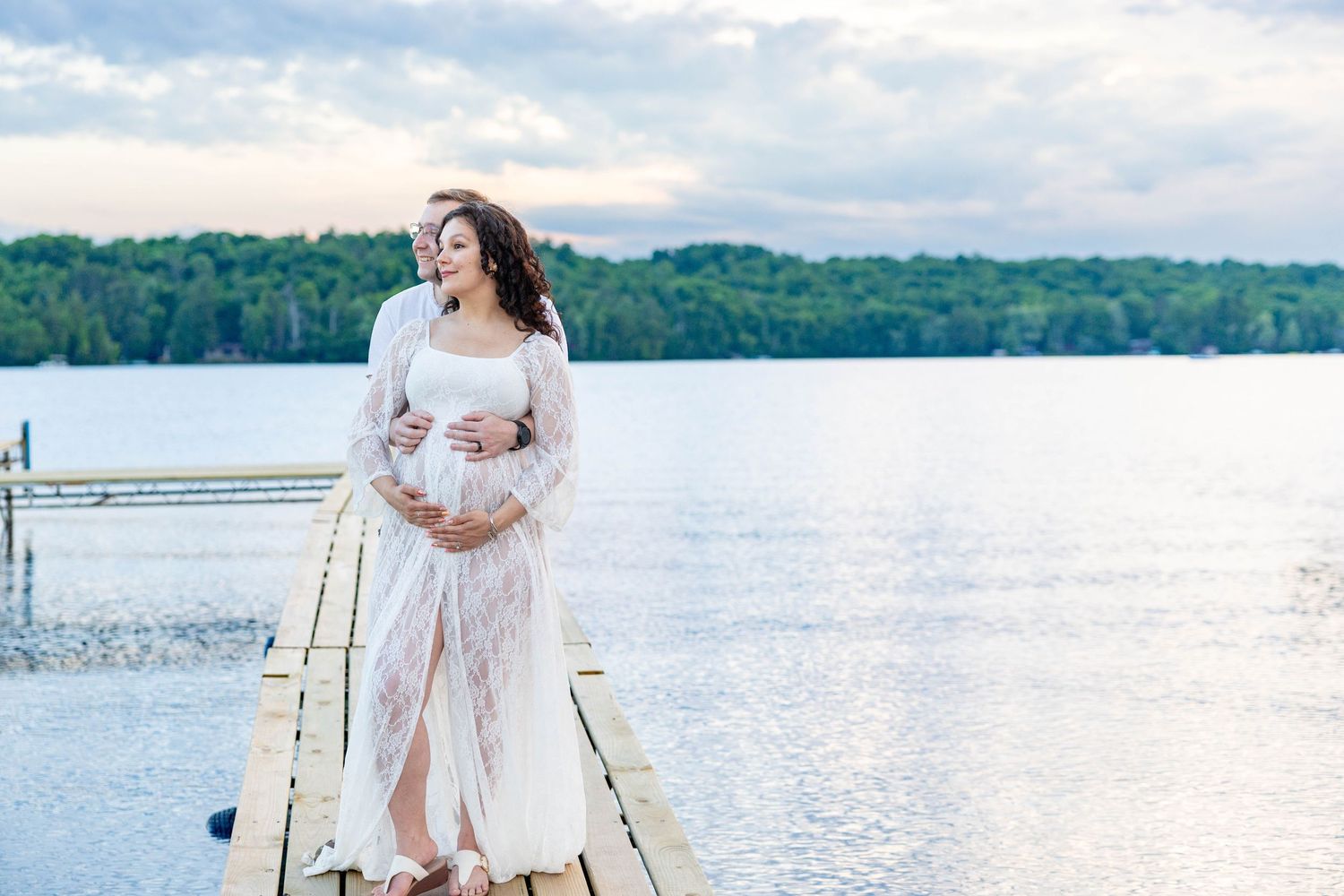 Pregnant woman in white lace gown standing on a dock by a serene lake, cradling her baby bump at sunset.