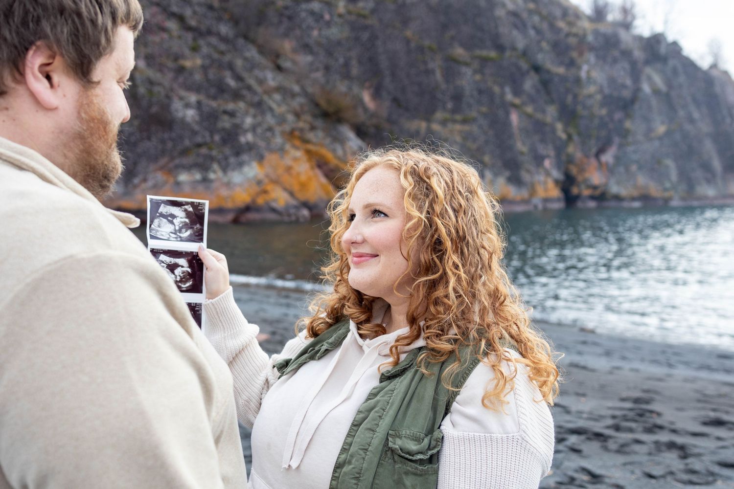 A couple shares a tender moment on a rocky beach while looking at an ultrasound photo.