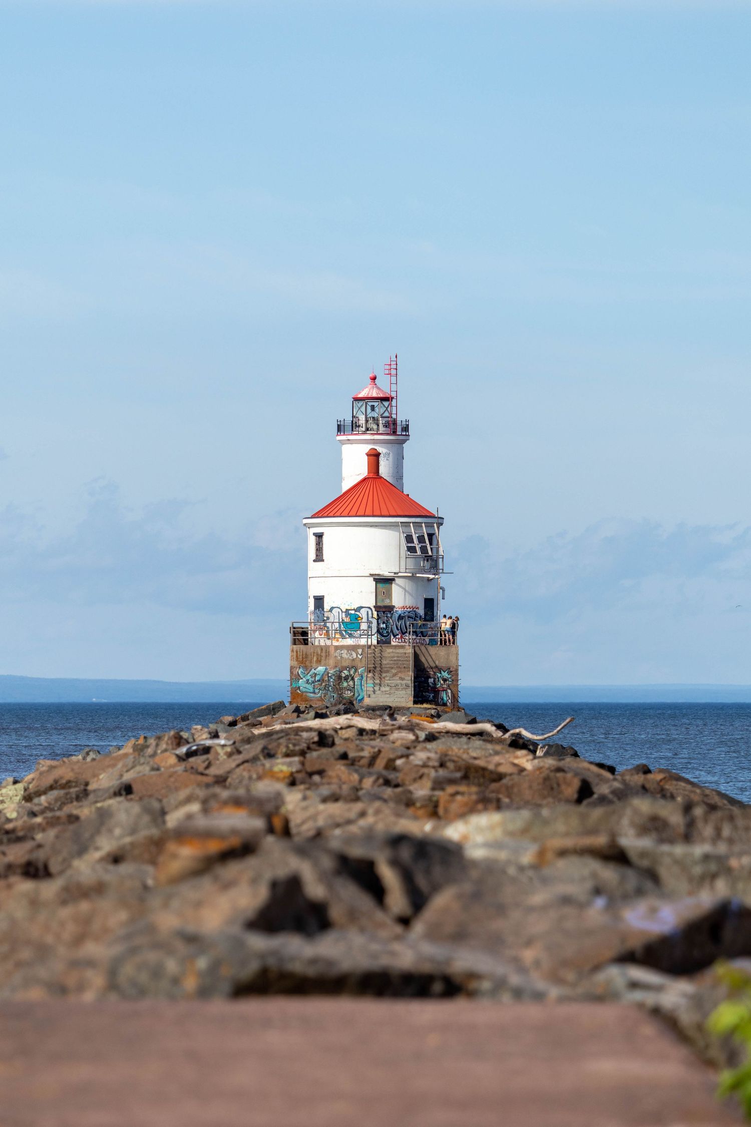 A small white lighthouse with red roof sits atop rocky coastal terrain against a blue ocean and sky background.