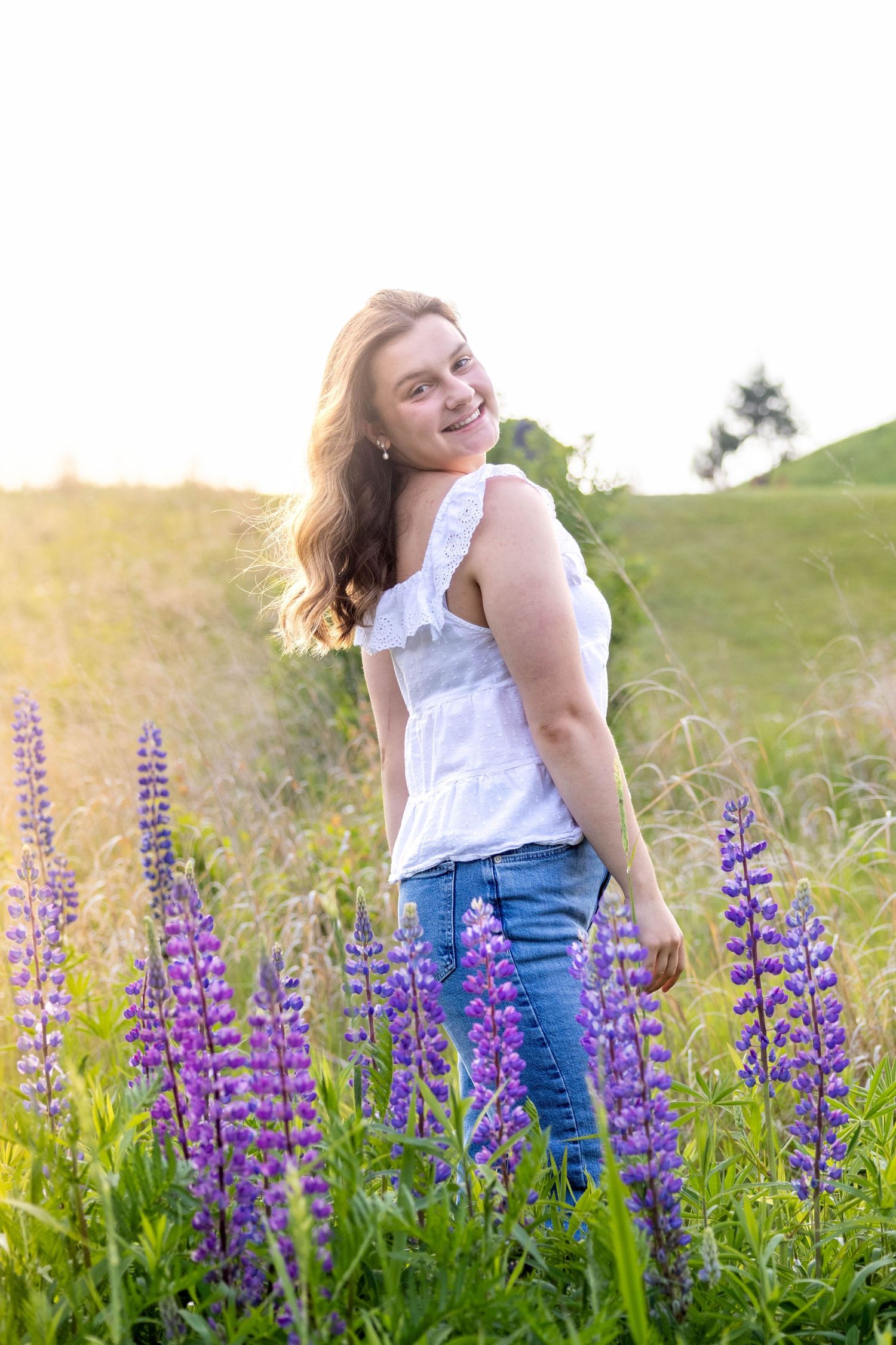 A person in a white top and jeans dances joyfully among vibrant purple lupine flowers in a sunlit meadow.